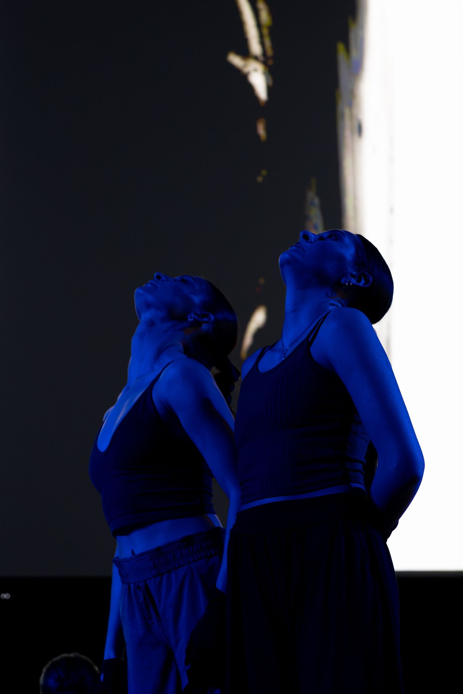 Two female dancers bathed in blue light perform with heads tilted back on stage.