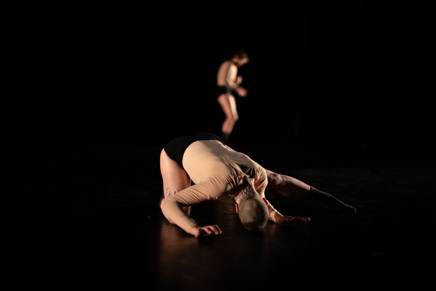 A contemporary dancer curls on a dark stage floor while another performer moves in the background.