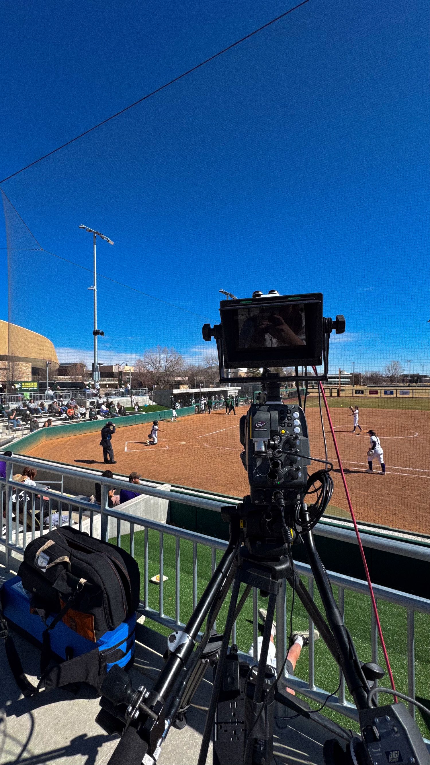 A professional video camera on tripod films a softball game at an outdoor stadium on a sunny day.
