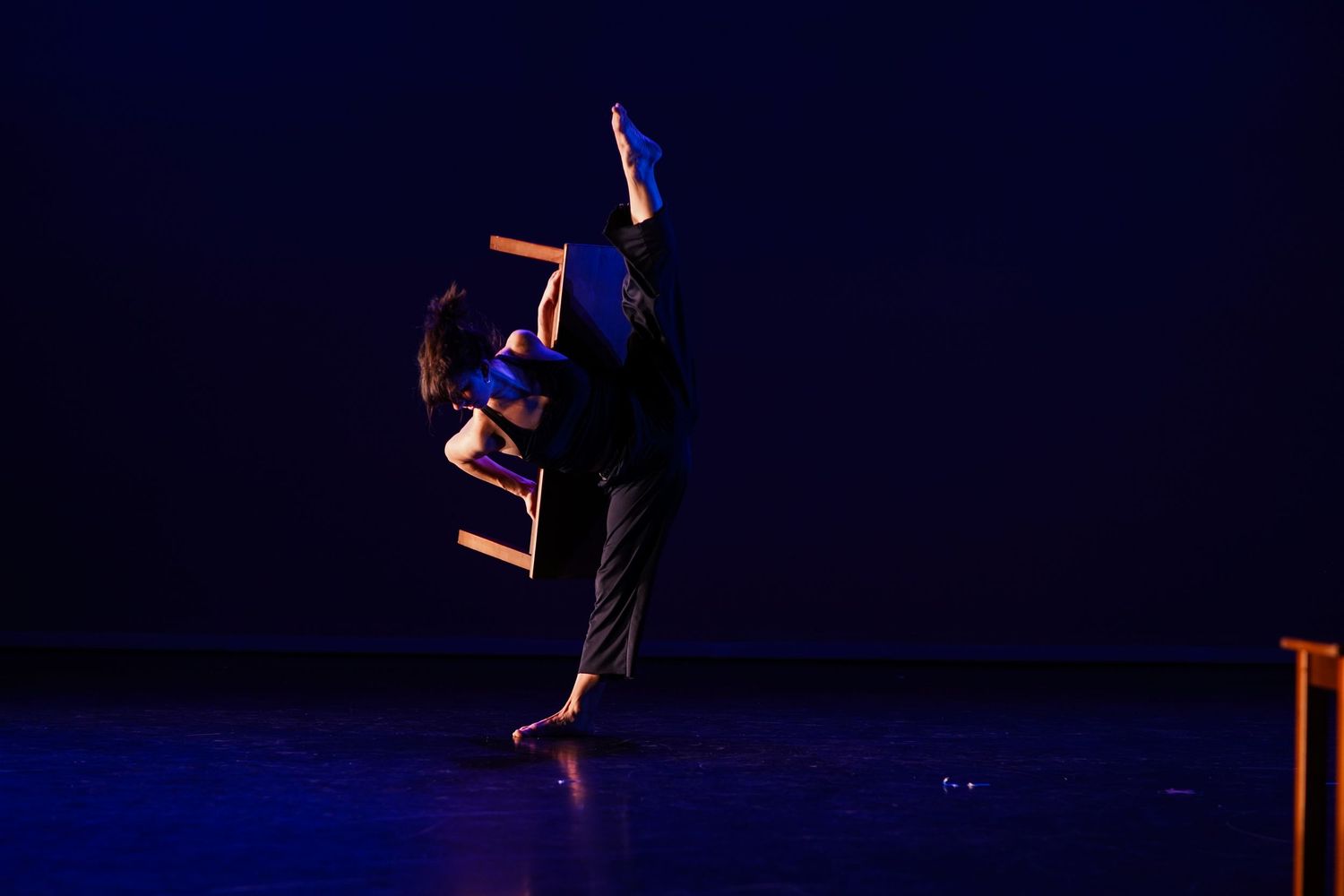 Solo contemporary dancer executes a high leg extension on a dark blue-lit stage in an expressive modern dance performance.