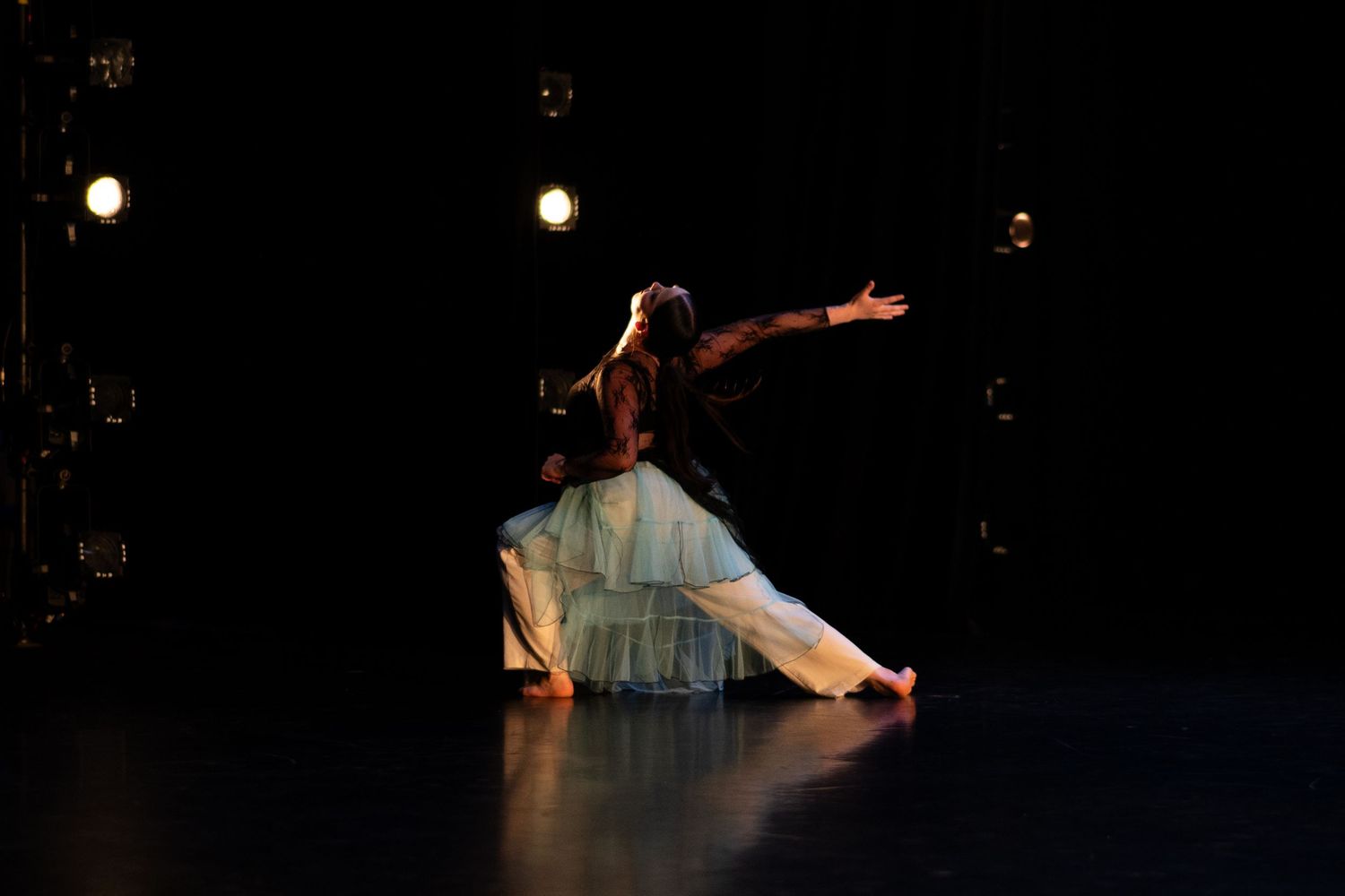 A dancer in a flowing white skirt performs an elegant pose on a dimly lit stage with spotlights.