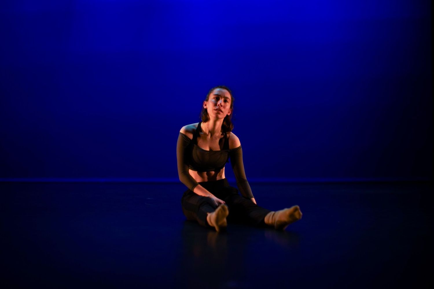A contemporary dancer sits on a dark stage floor bathed in vivid blue light during a solo performance.