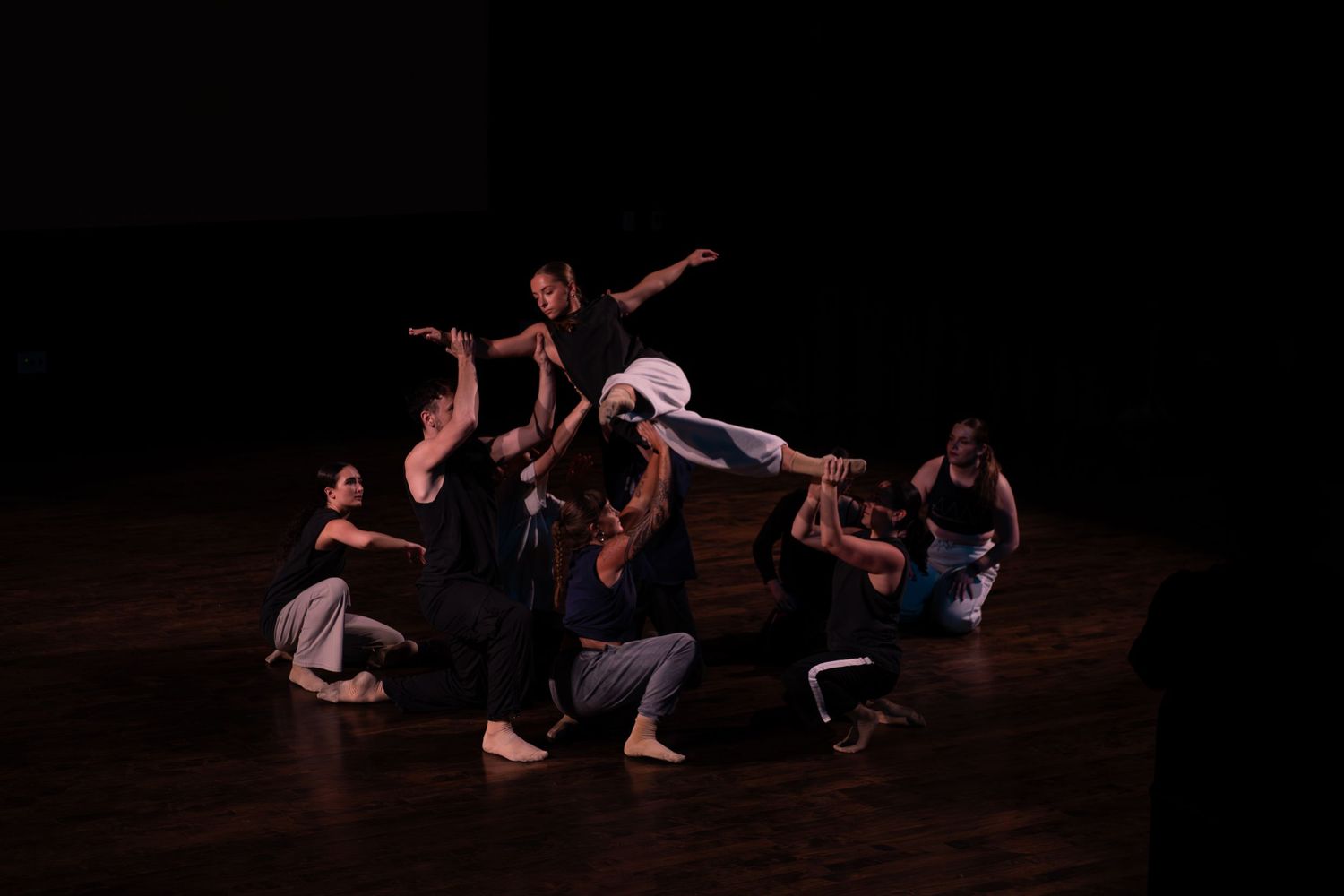 A group of contemporary dancers perform a dramatic ensemble piece on a dimly lit theater stage.