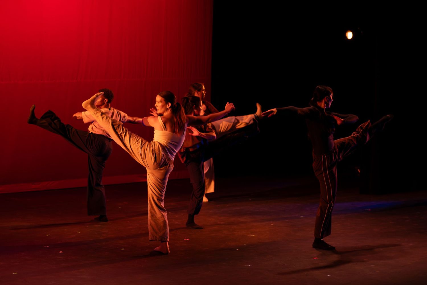 Contemporary dancers execute high kicks in synchronized choreography under red stage lighting.