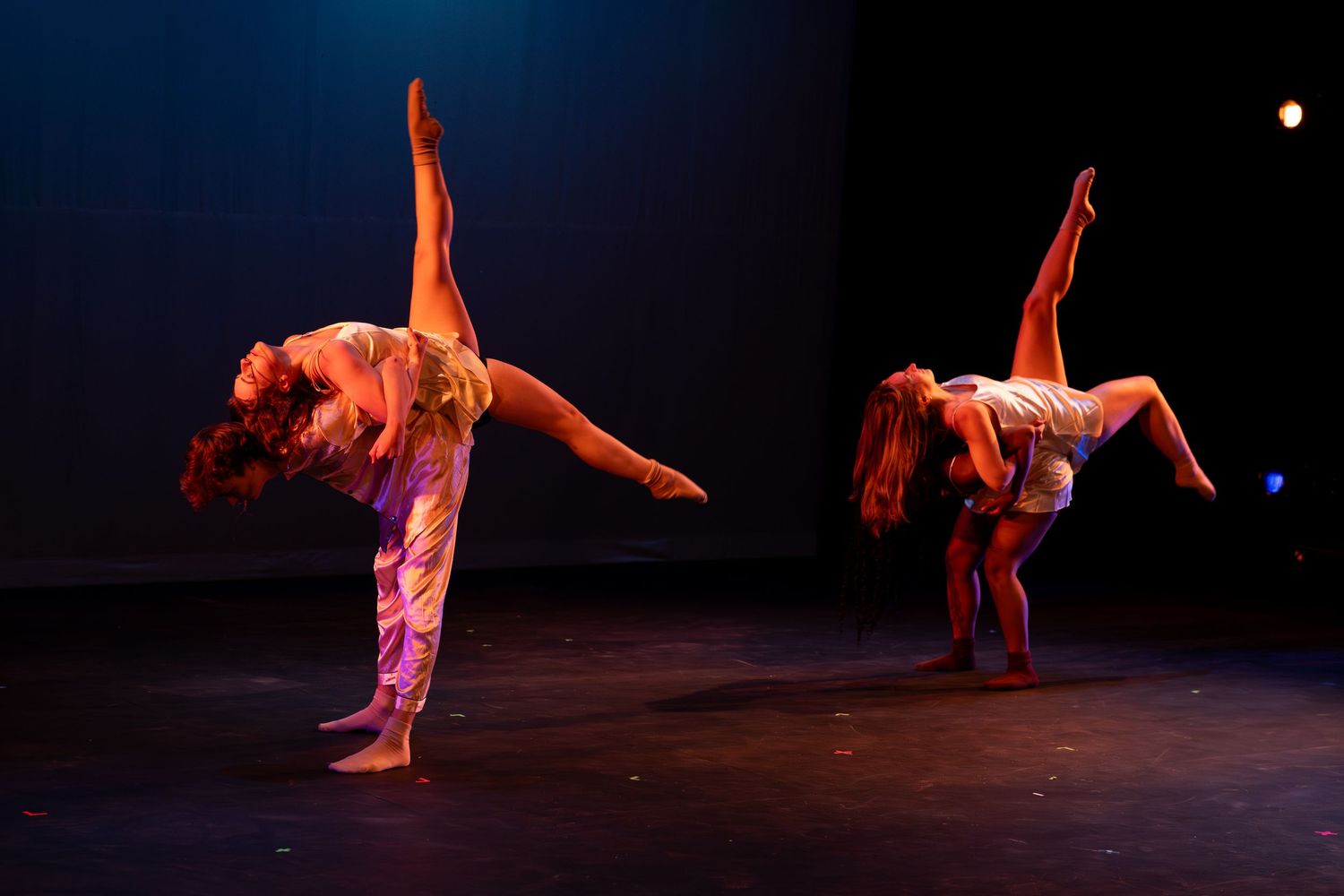 Two dancers execute a synchronized arabesque balance pose on a dimly lit performance stage.