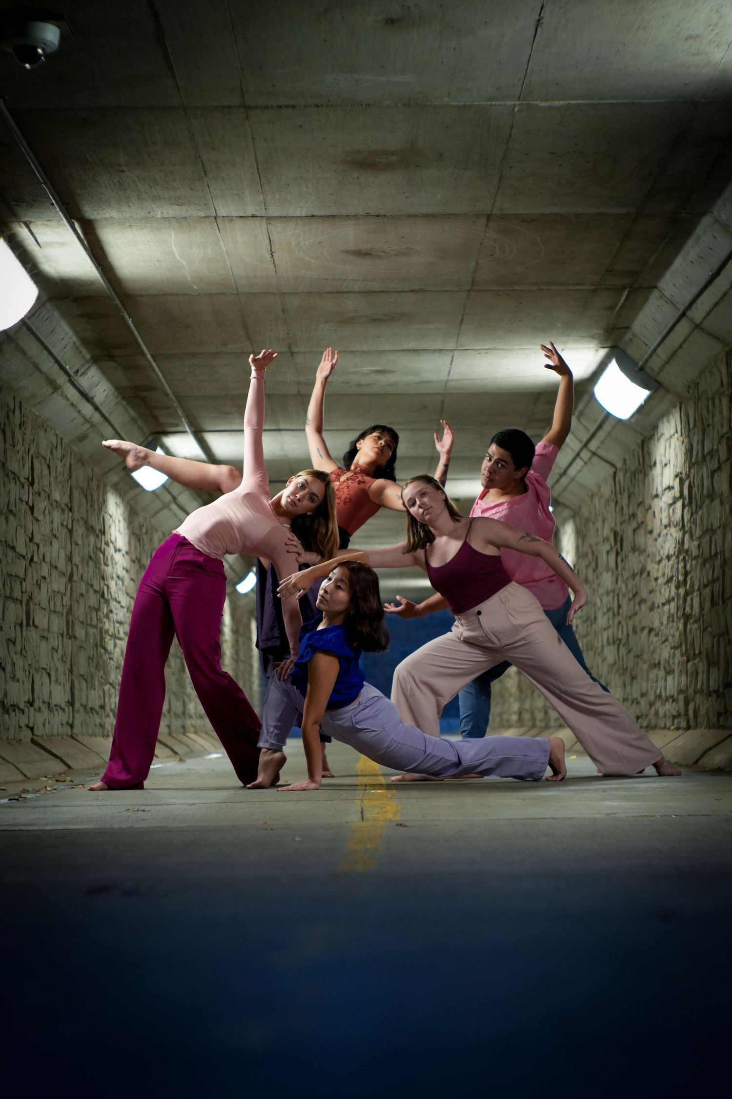 Five dancers strike dynamic poses in a concrete tunnel, wearing colorful attire under fluorescent lights.