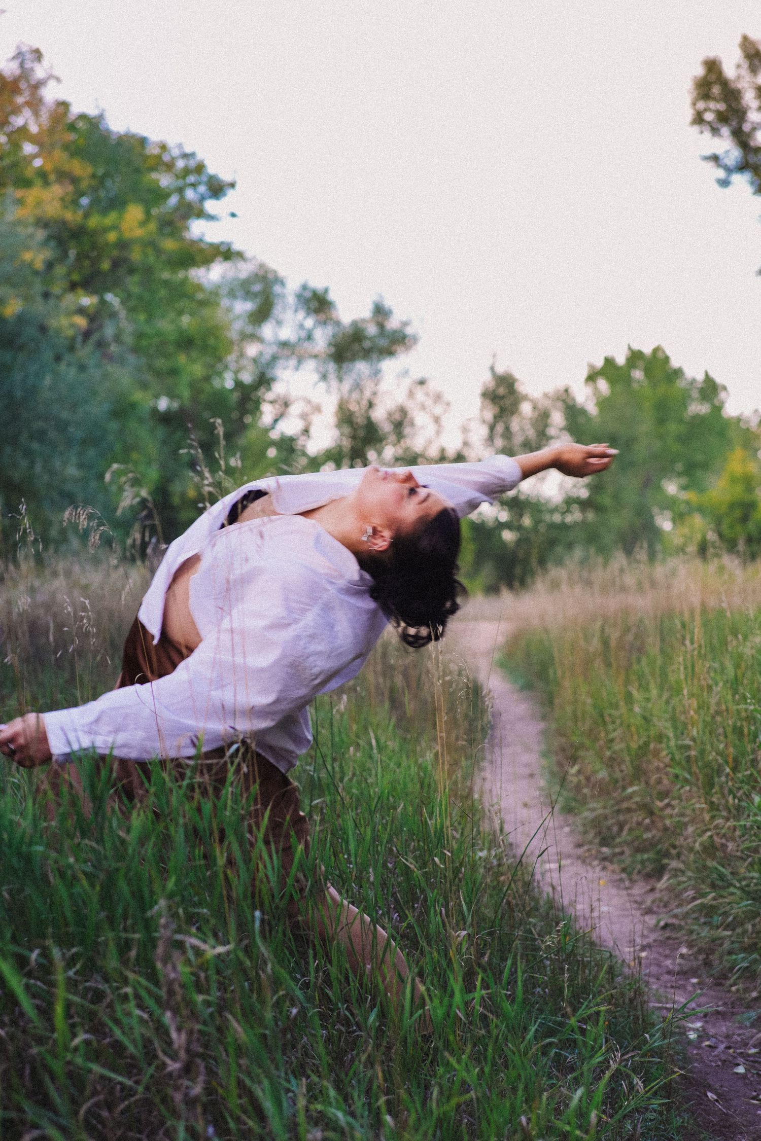 A dancer in a white shirt performs a dramatic backbend on a lush green nature trail at dusk.
