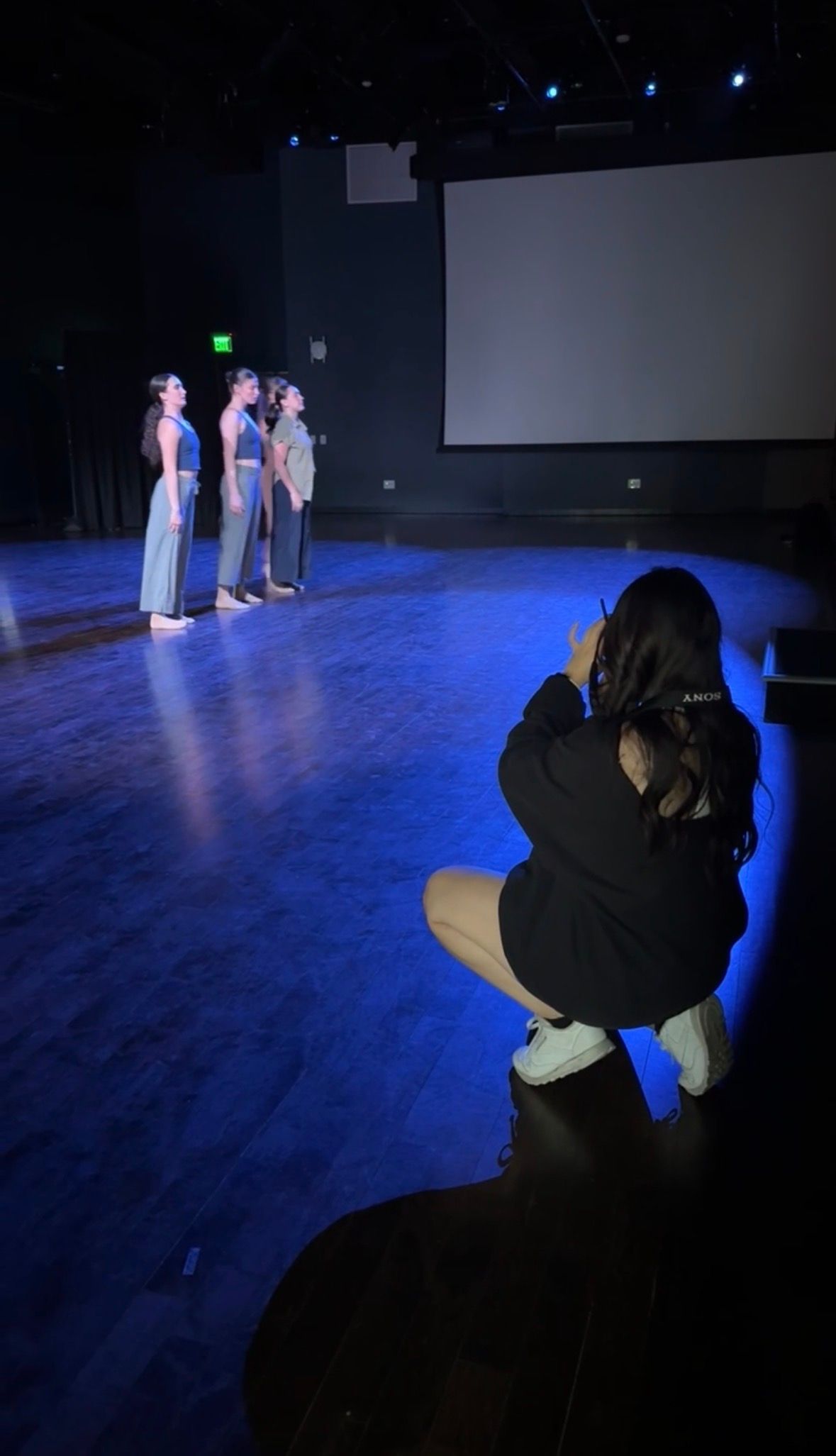 Photographer crouches to capture three dancers in gray costumes on blue-lit stage.