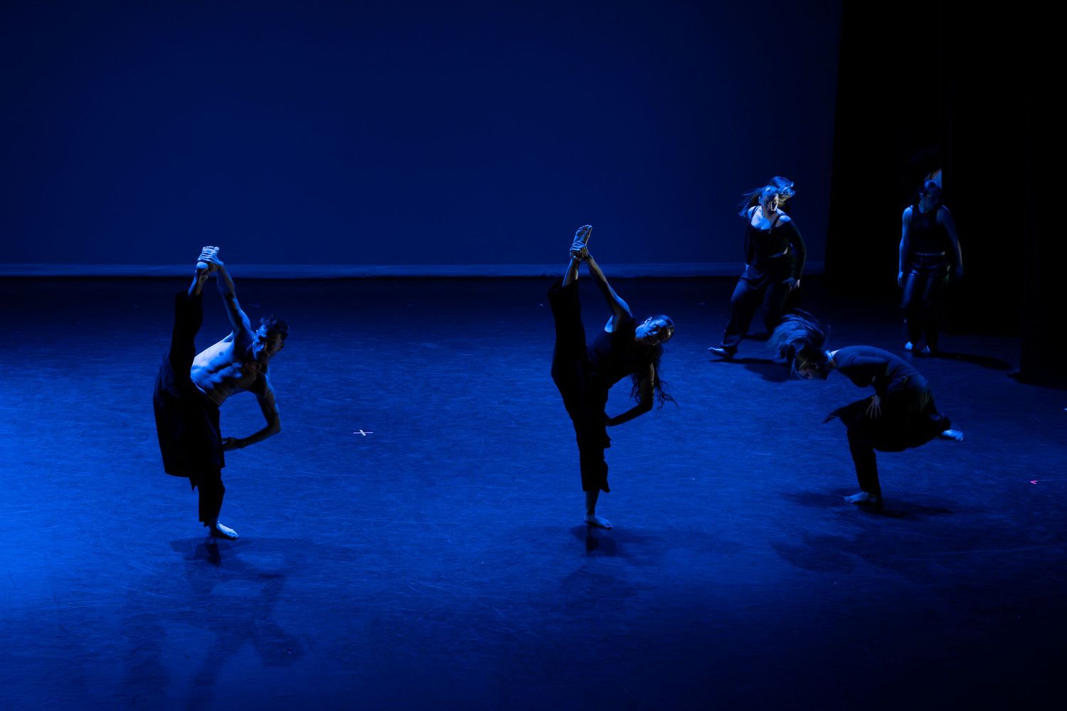 Four dancers in black execute high leg extensions in a contemporary dance performance under blue stage lighting.