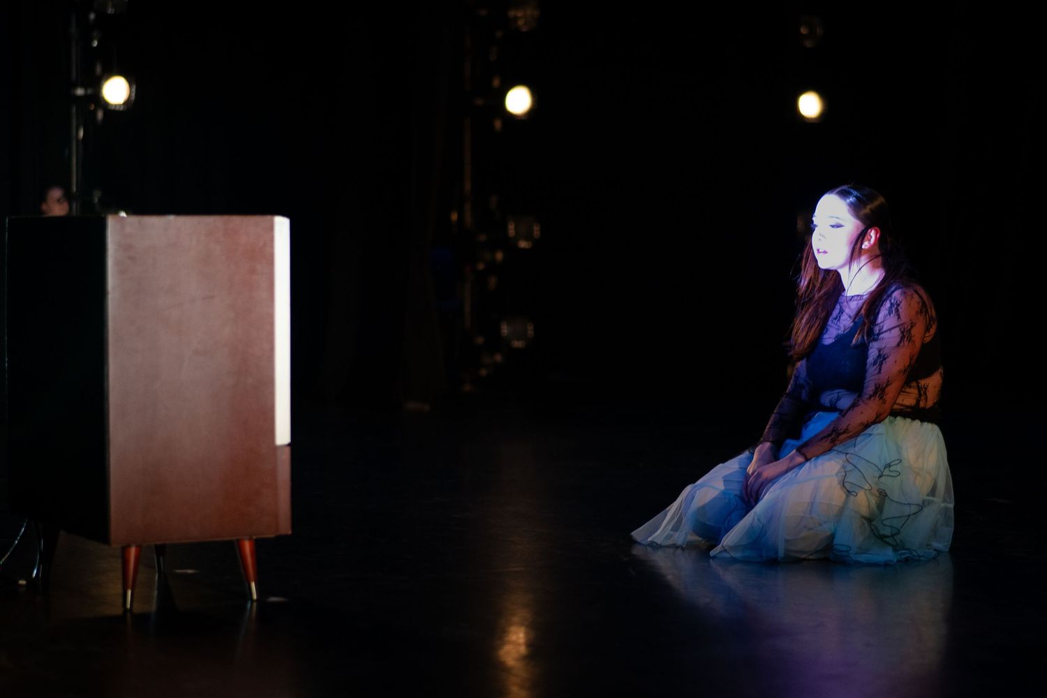 Female dancer sits on dark stage beside a vintage wooden cabinet under spotlight.