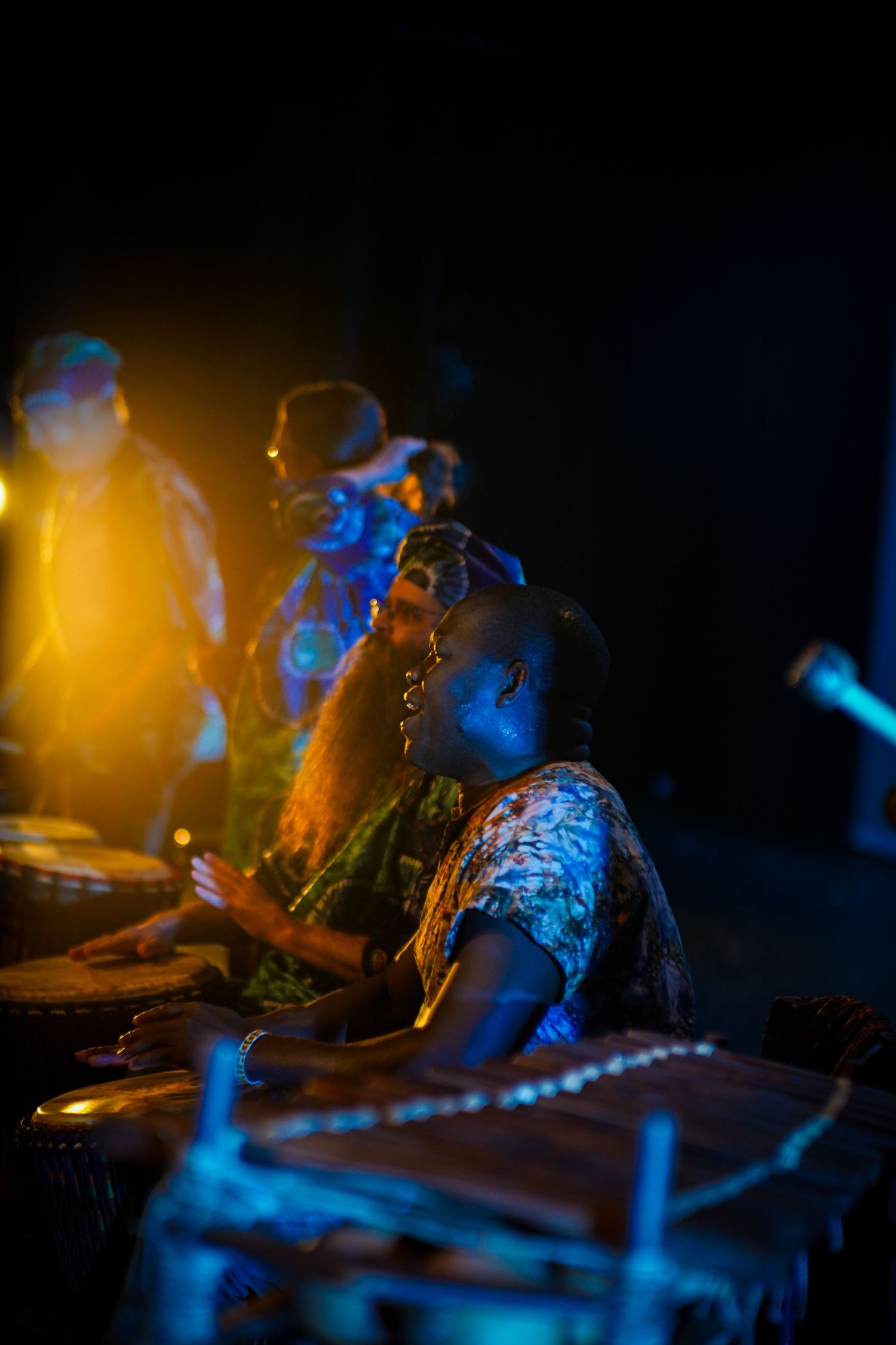Musicians play traditional African djembe drums on stage under dramatic warm golden and blue stage lighting.