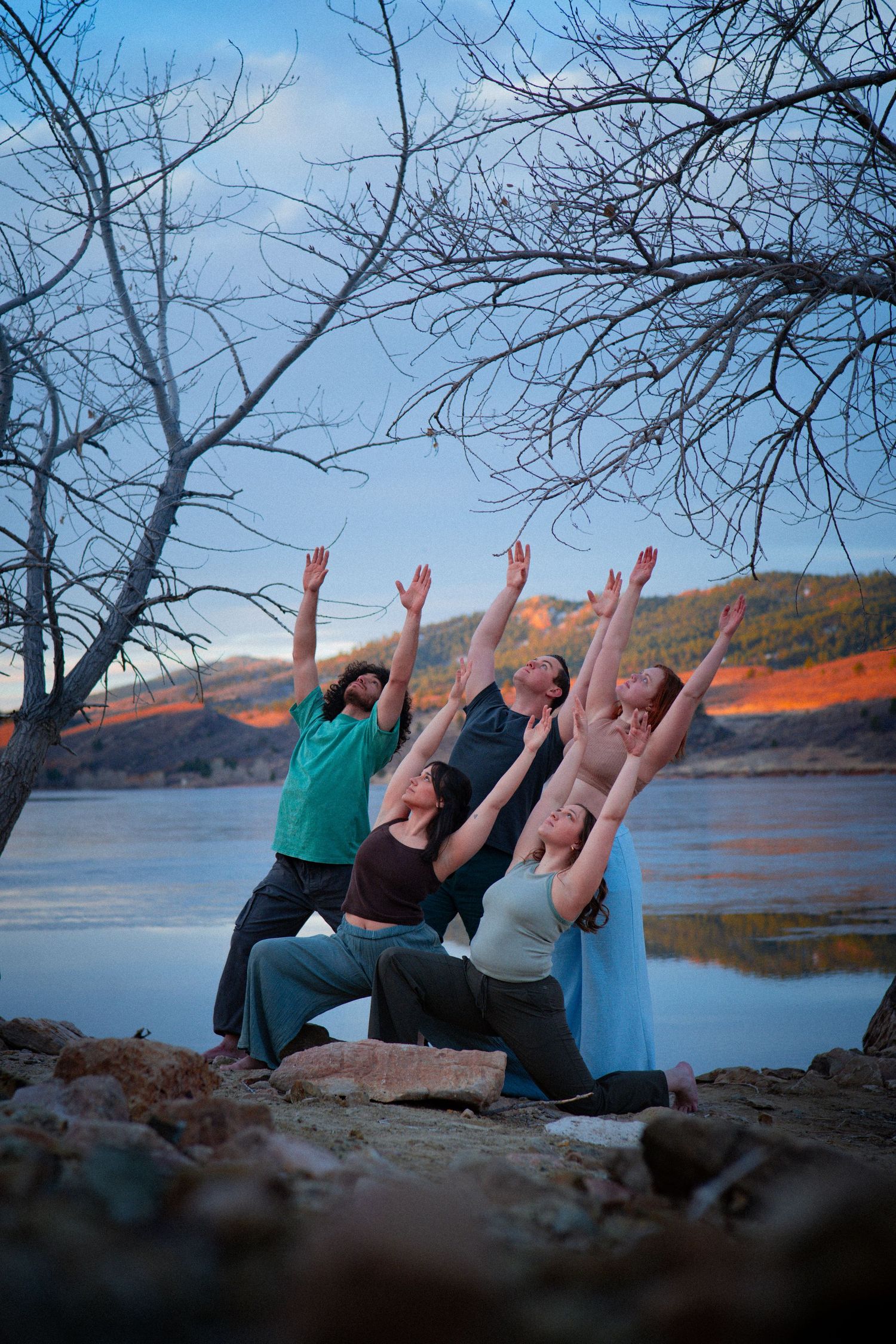 Four dancers kneel with arms raised toward sky near a lakeside with bare trees at dusk.