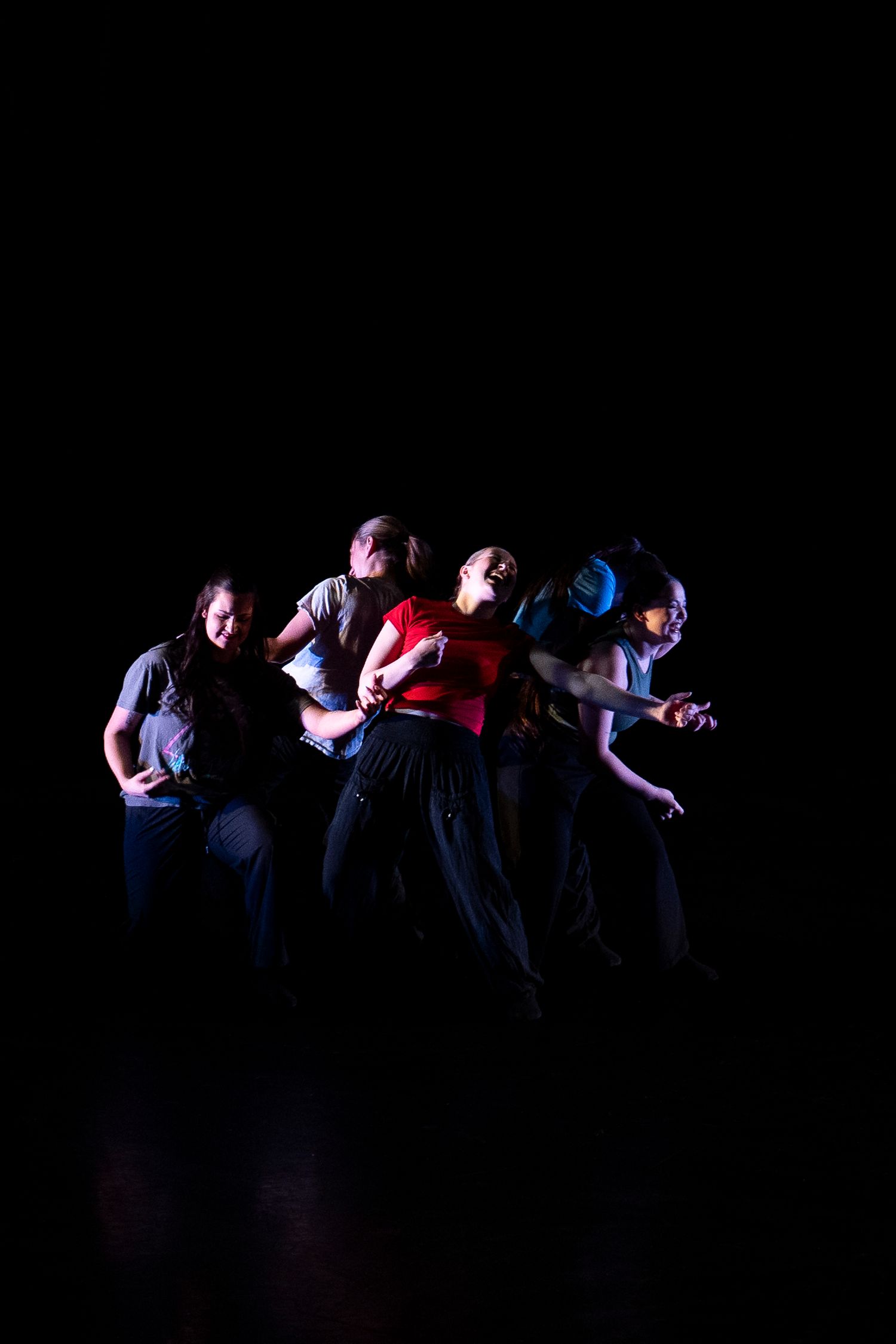 Five contemporary dancers perform an expressive group routine under colorful stage lighting against a black backdrop.