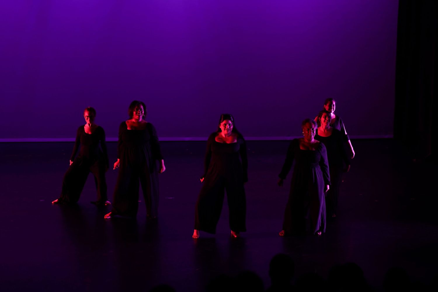 Four contemporary dancers in black costumes perform on stage under striking purple stage lighting.