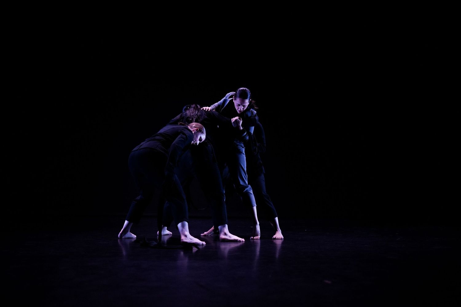 Three dancers in black clothing perform a huddled contemporary dance piece on a dark stage.
