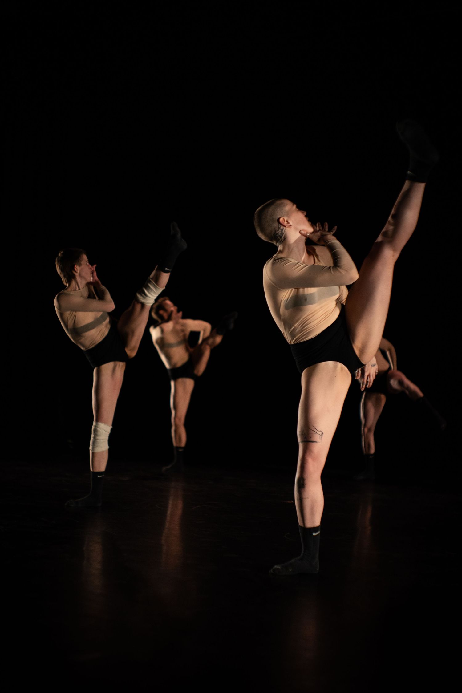 Contemporary dancers in nude bodysuits perform high leg extensions on a dark stage during a group performance.