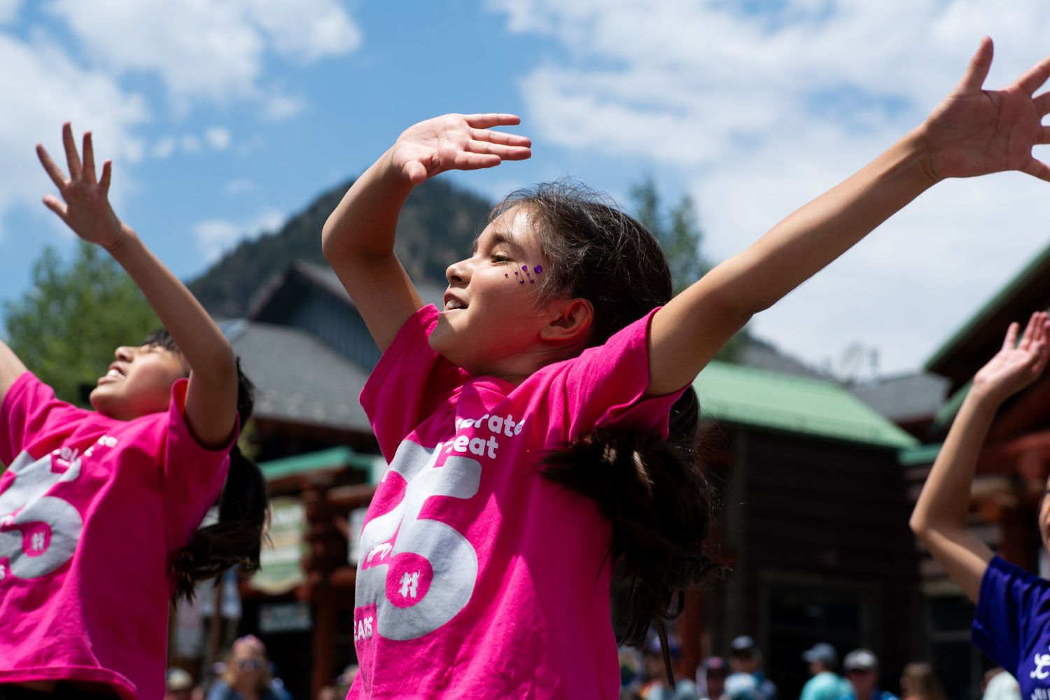 A girl in a pink t-shirt joyfully raises her arms skyward during an outdoor summer camp activity.