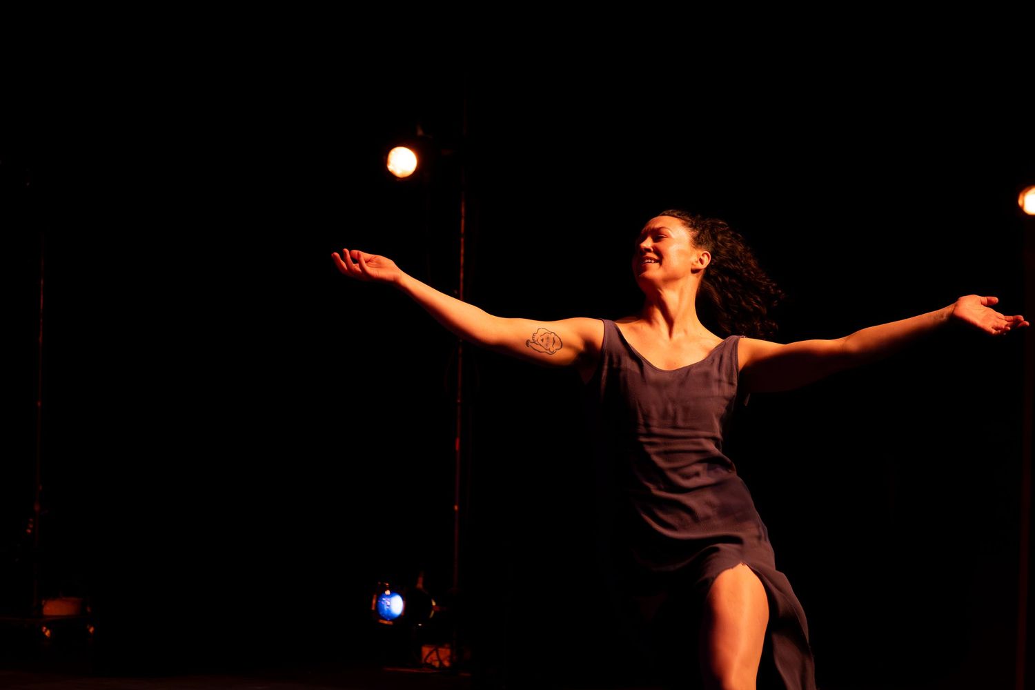 Female dancer extends arms wide in a graceful pose under warm spotlight on a dark stage.