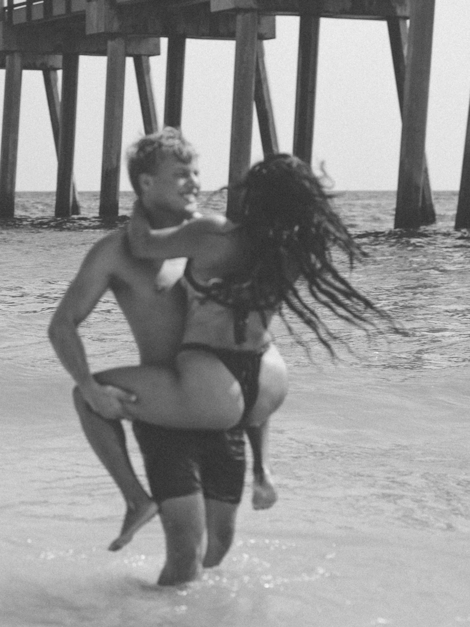 Black and white photo of a man carrying his partner under a fishing pier