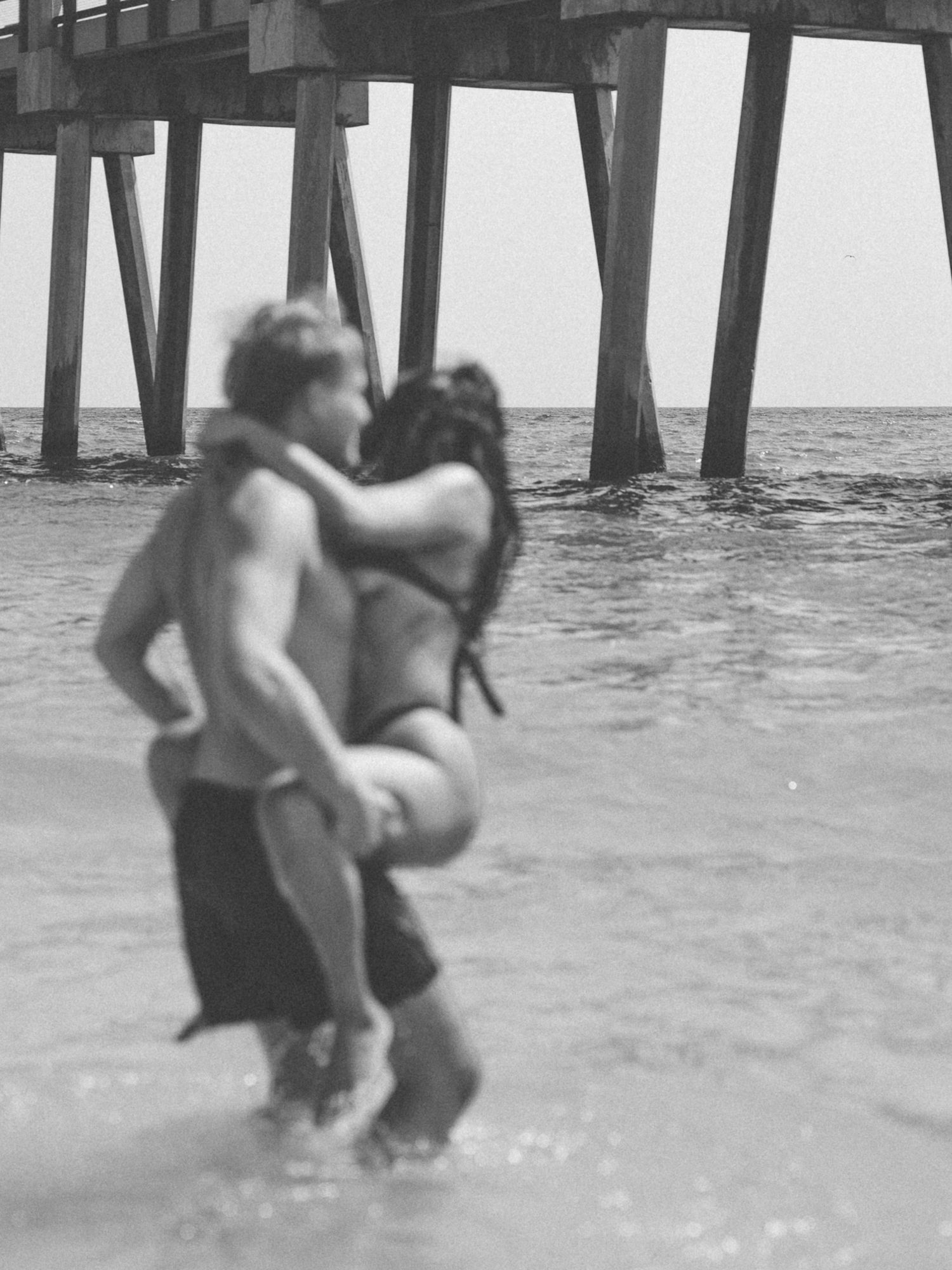Black and white photo of a couple embracing in the water under a fishing pier