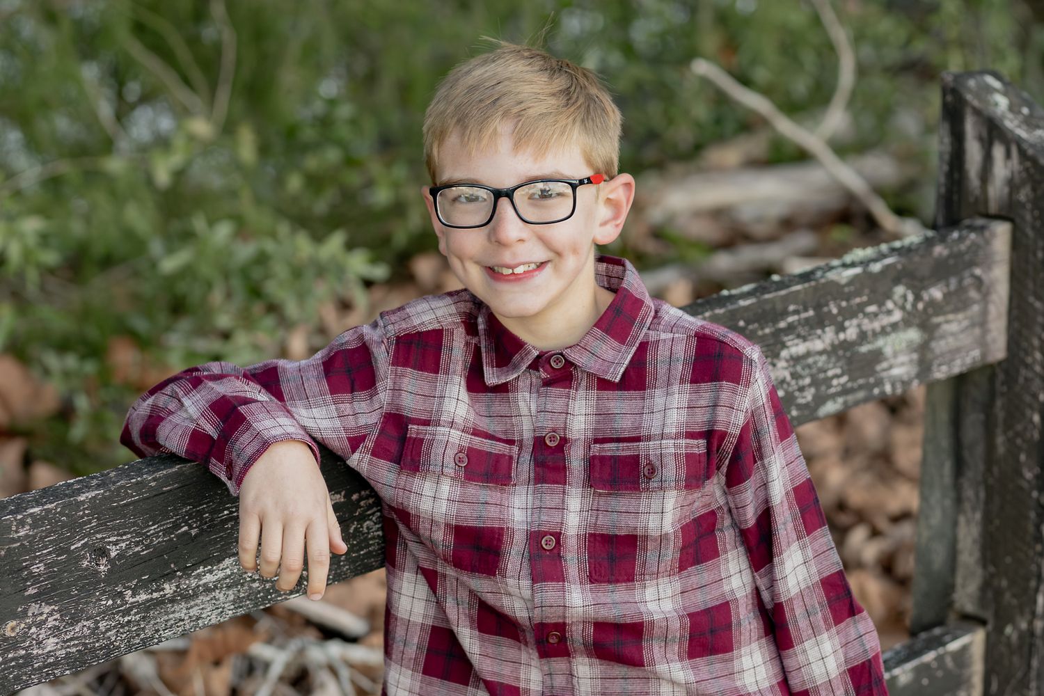 Boy in glasses and a plaid shirt leaning on a wooden fence