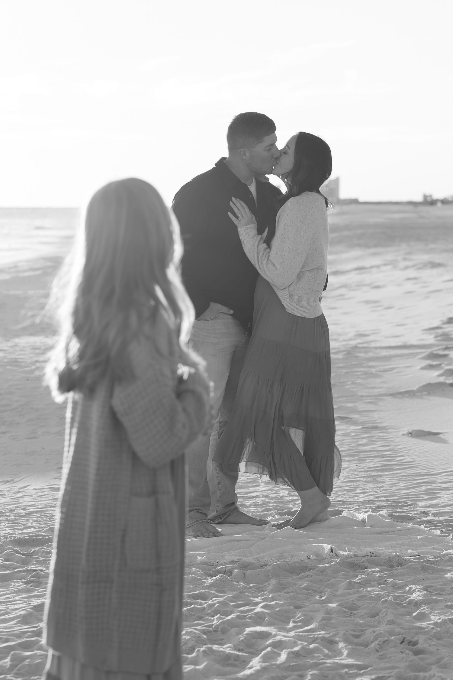 Black and white photo of a couple kissing on the beach while their daughter looks on