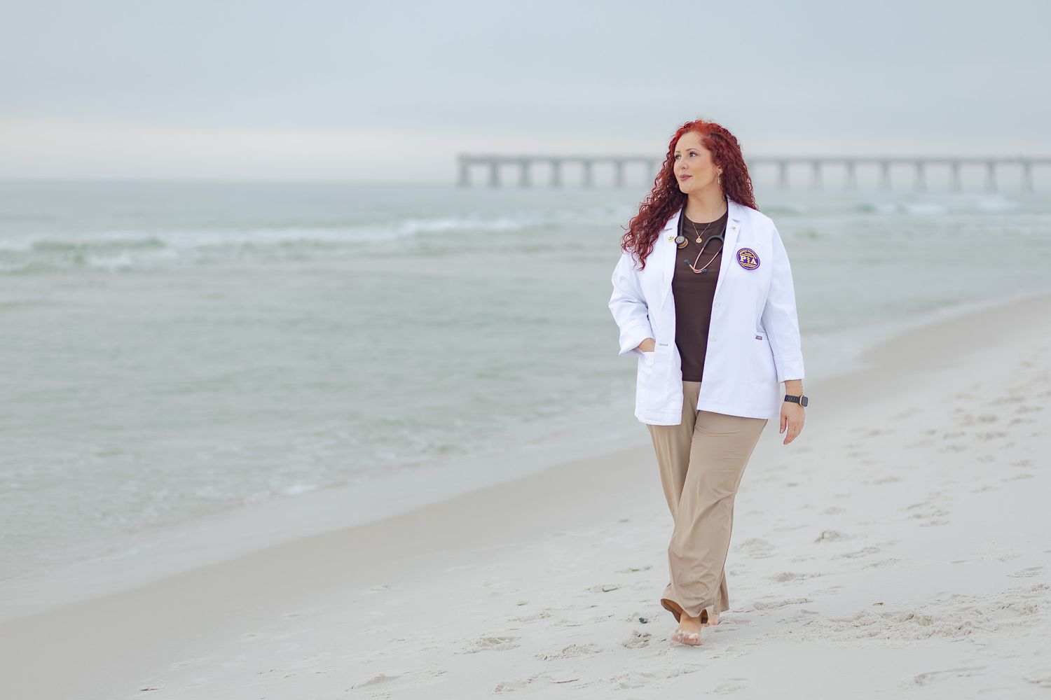 Graduate in her white coat walking the beach with a pier in the distance