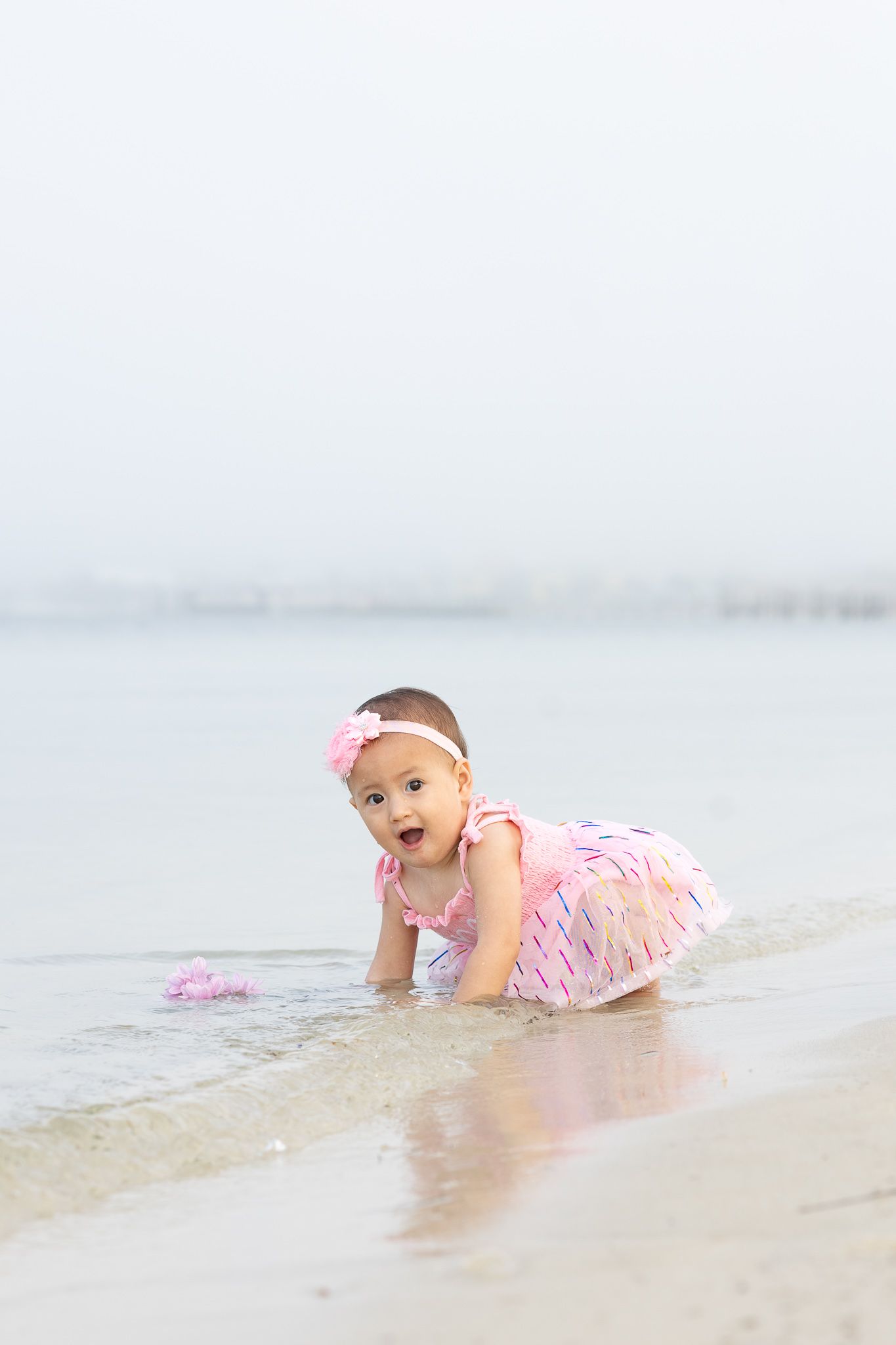 Baby girl in a pink dress crawling at the water's edge, first birthday beach session