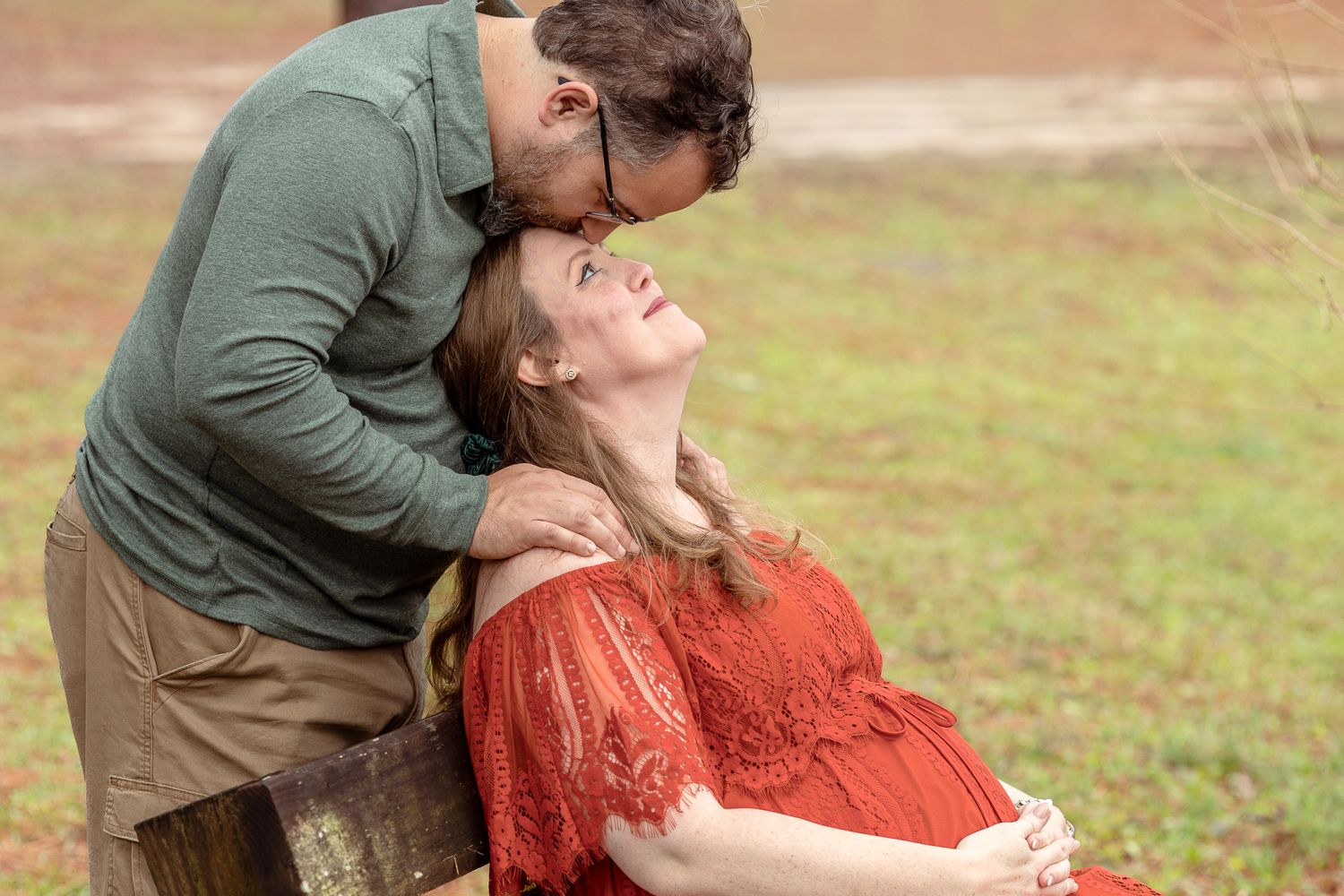 Husband kissing his pregnant wife's forehead in a red lace dress at Bear Lake, Milton FL