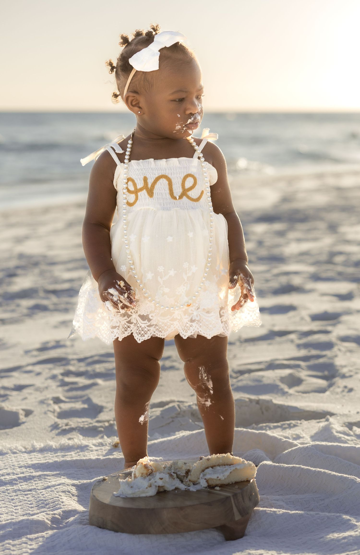 Baby girl in a white 'one' romper standing on a wood slab on the beach