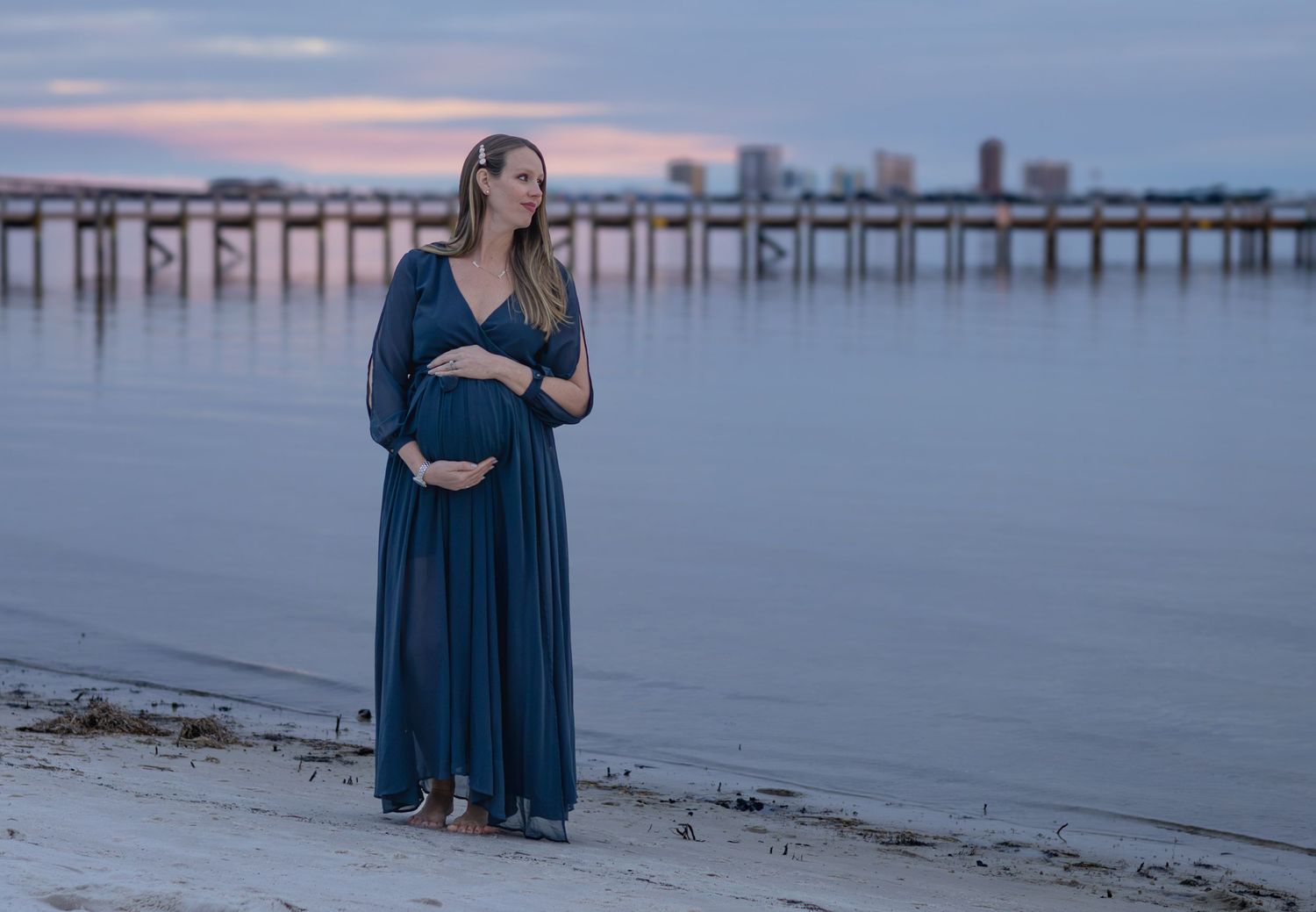 Expecting mother in a navy dress on the shore at dusk with a pier behind her