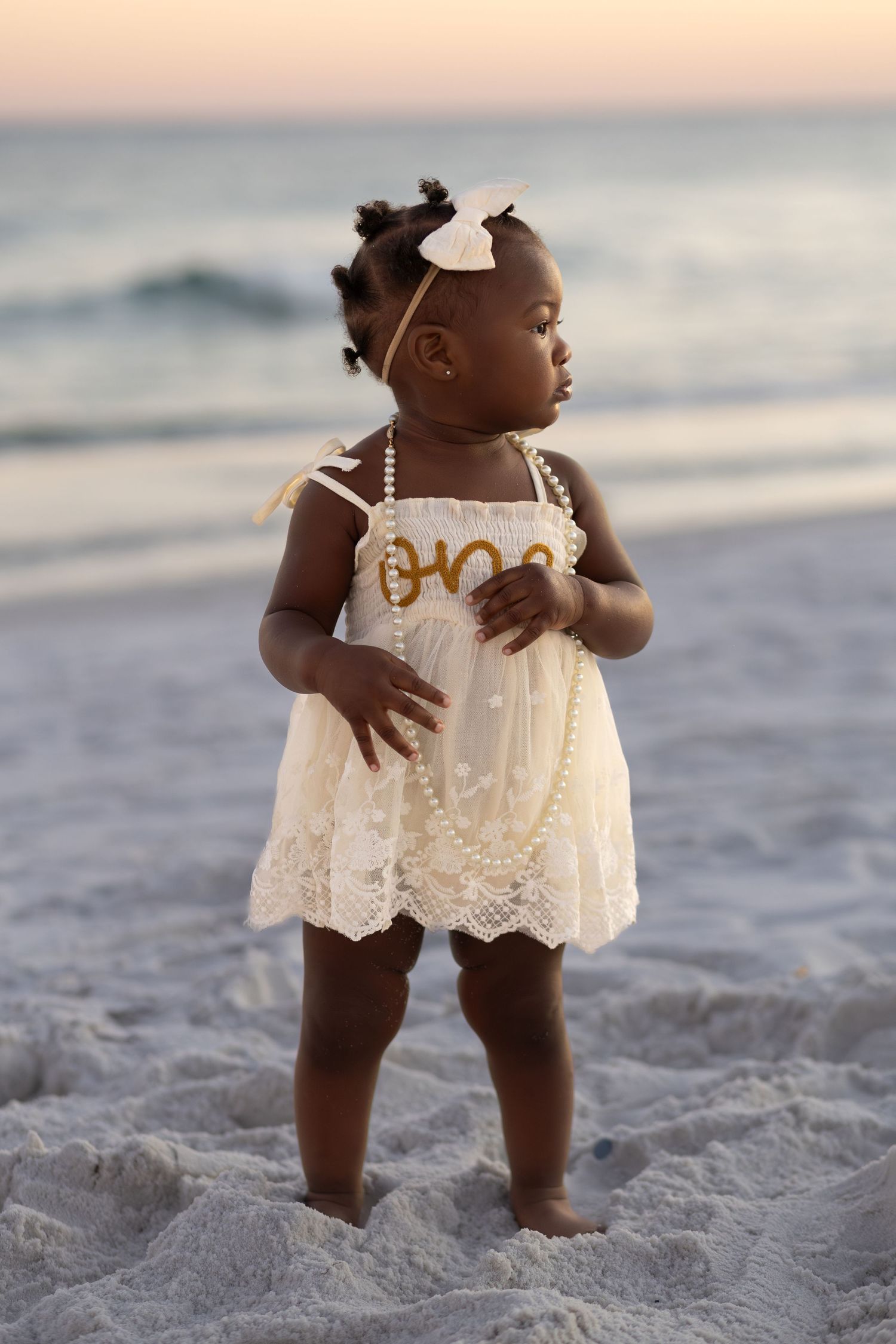 Baby girl in a 'one' outfit standing on the beach at sunset