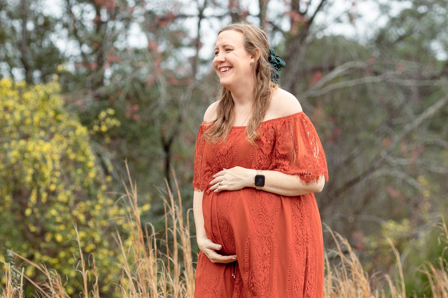 Smiling mom-to-be in a red maternity dress among the trees at Bear Lake, Milton FL
