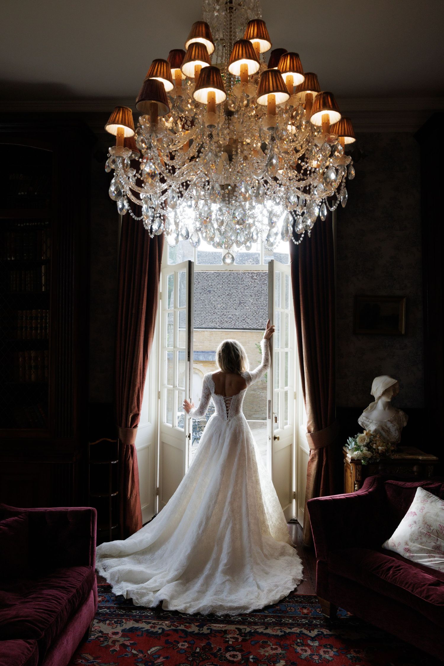 Bride in a stunning lace wedding gown standing beneath a crystal chandelier gazing out ornate open French doors.