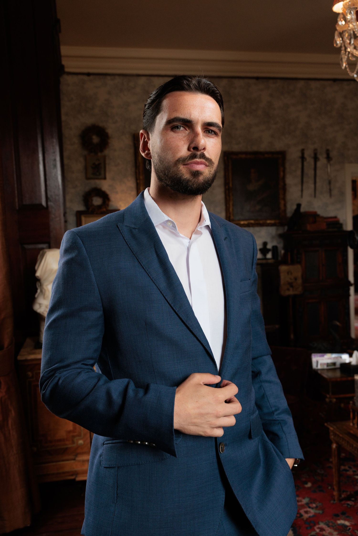 Handsome groom in a navy blue suit adjusting his jacket in an elegantly decorated vintage interior room.