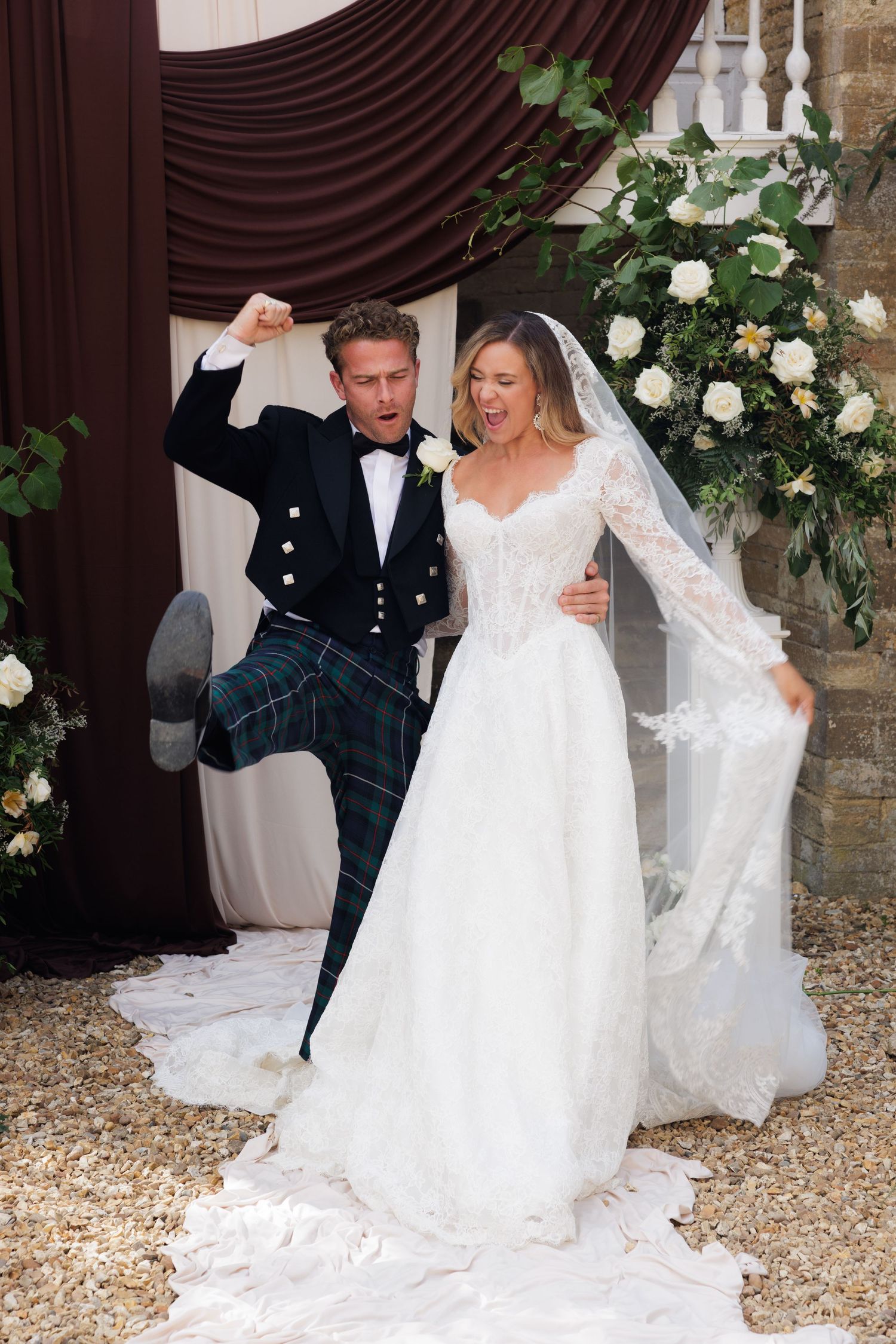 Jubilant groom in tartan kilt celebrates with bride in lace wedding gown at floral ceremony arch.
Greater Manchester