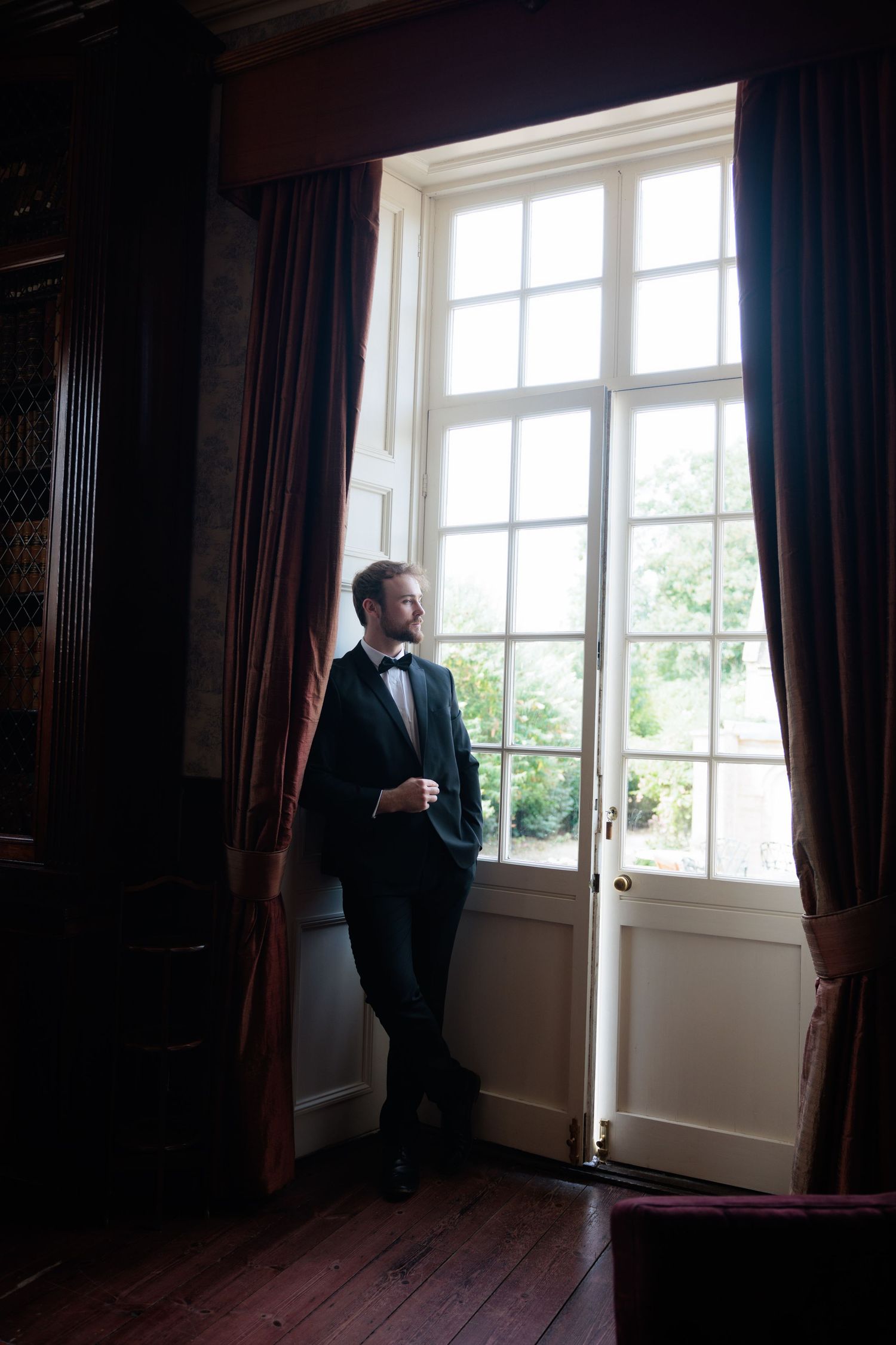 Groom in a black tuxedo gazing thoughtfully out a tall French window with red velvet curtains in a grand room.