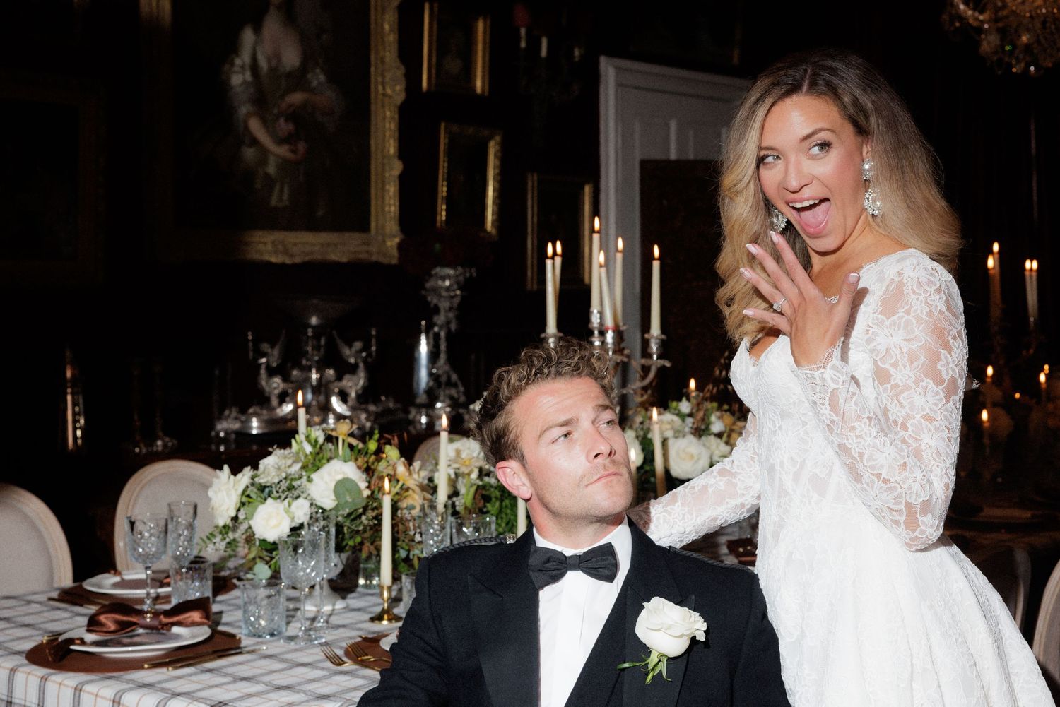 Bride in lace wedding gown laughing with groom in tuxedo at elegant candlelit wedding reception dinner table.