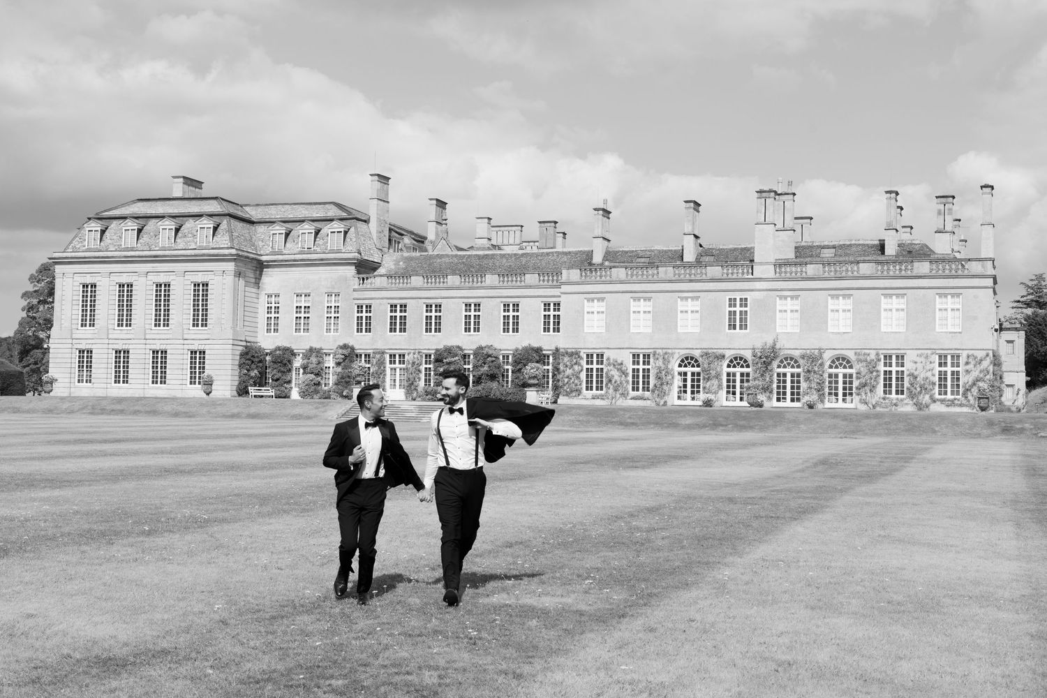 Two grooms in tuxedos walk across grand estate lawn in elegant black and white wedding portrait.