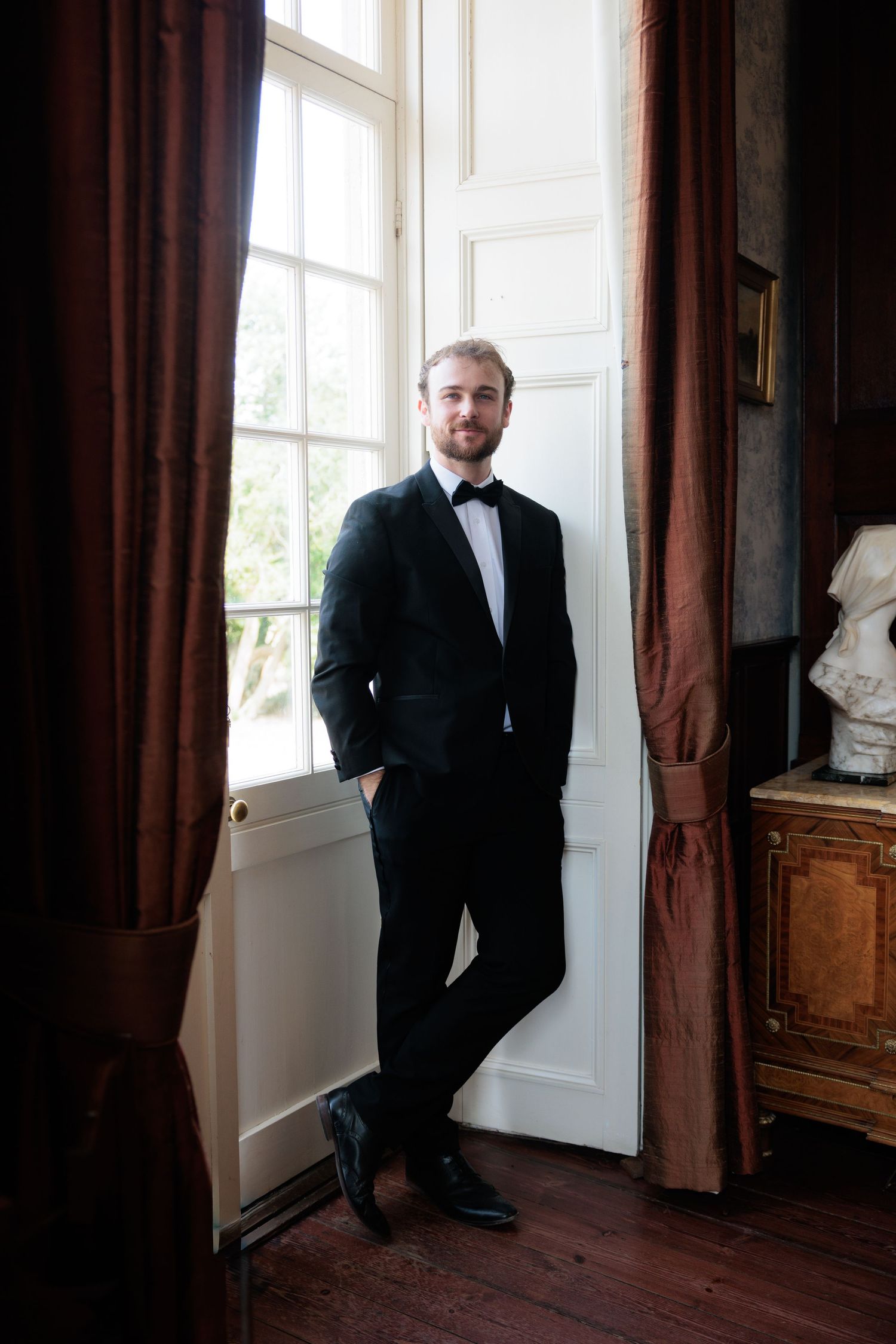 Groom in a black tuxedo with bow tie standing confidently by a tall window with red curtains indoors.