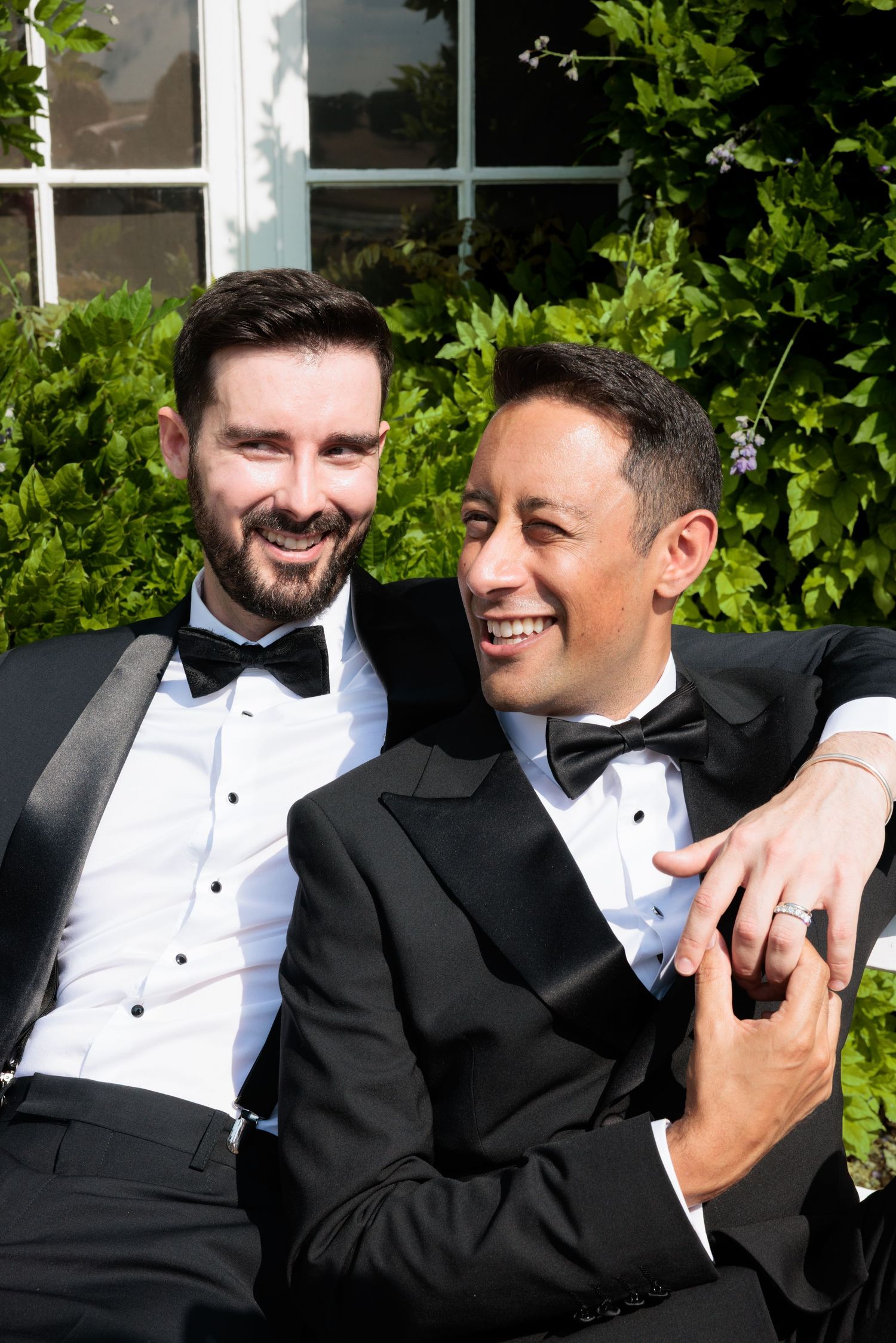 Two grooms in black tuxedos with bow ties laughing and embracing while seated outdoors surrounded by greenery.