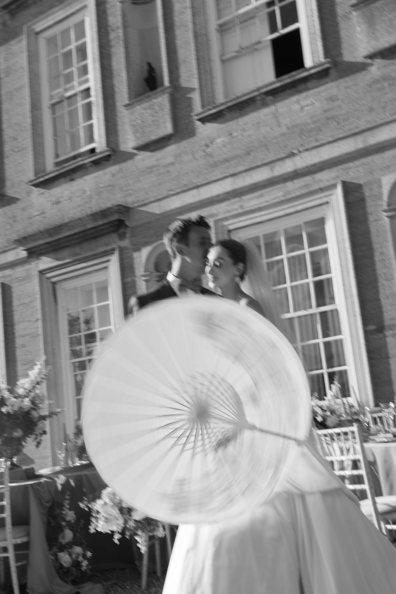 Blurred black and white photo of newlyweds kissing with parasol outside grand country house.