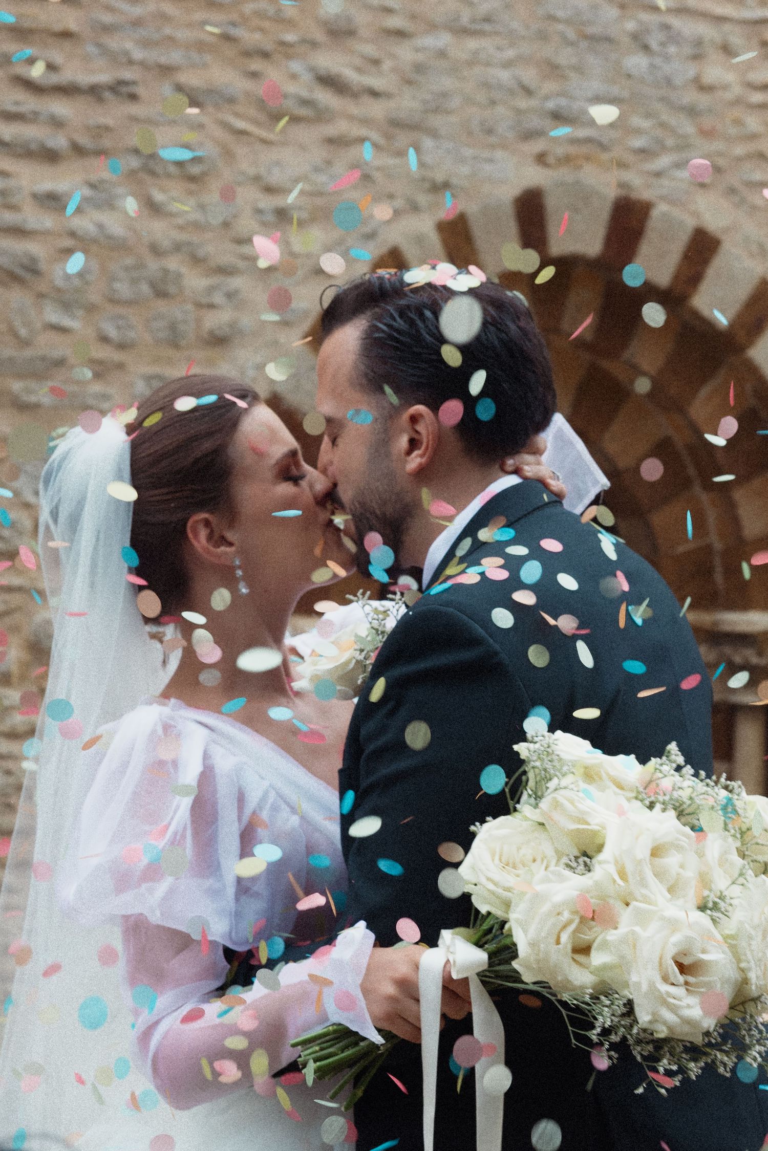 Newlyweds share first kiss surrounded by colorful confetti outside rustic stone church.