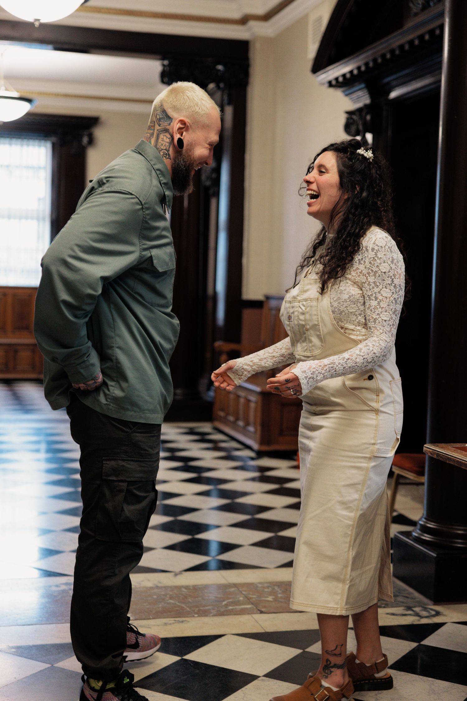 Laughing bride and tattooed groom share joyful moment on black and white checkered floor at wedding venue.