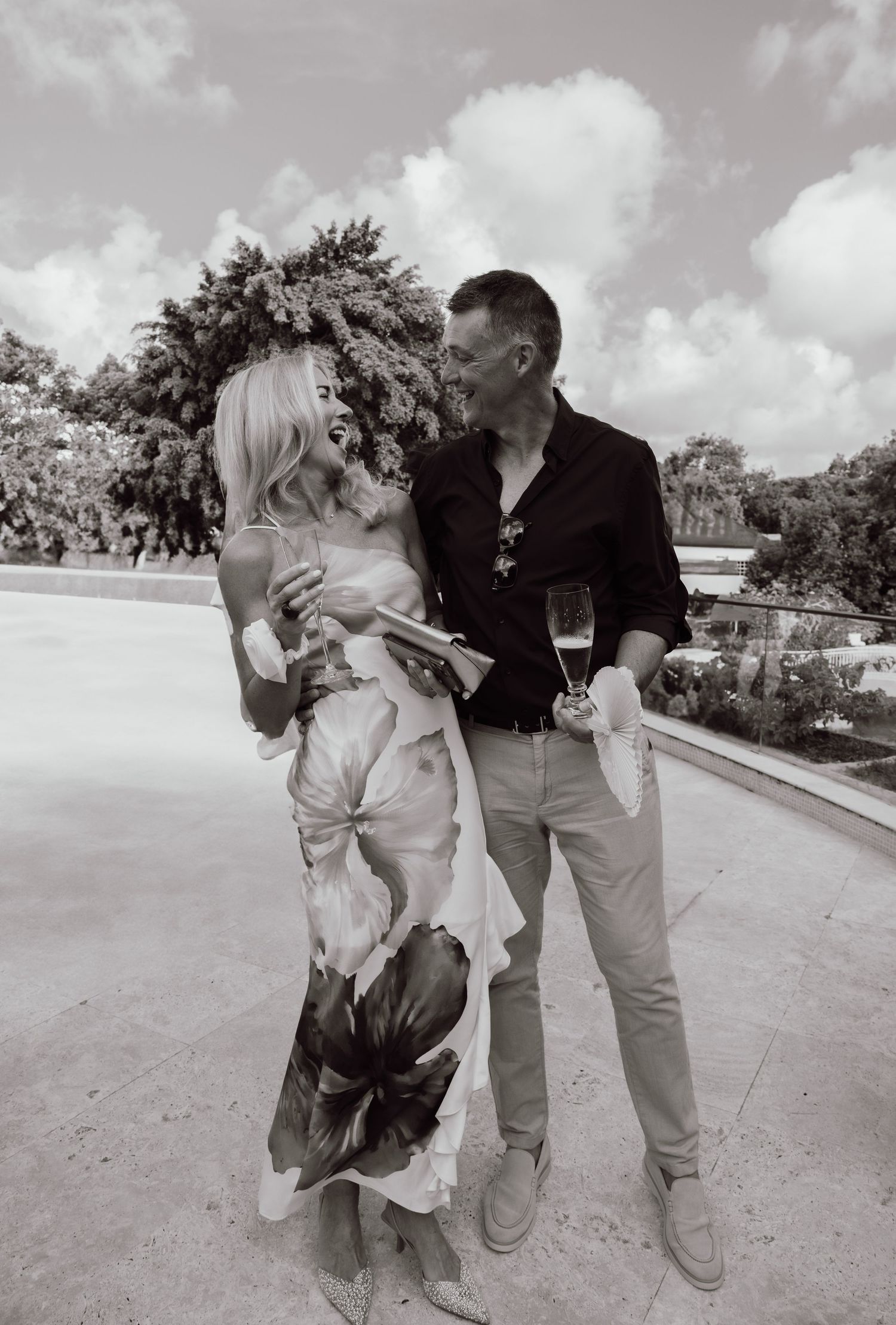 Elegant couple sharing a romantic moment outdoors, holding champagne glasses, dressed in floral dress and black shirt. Barbados Hotel