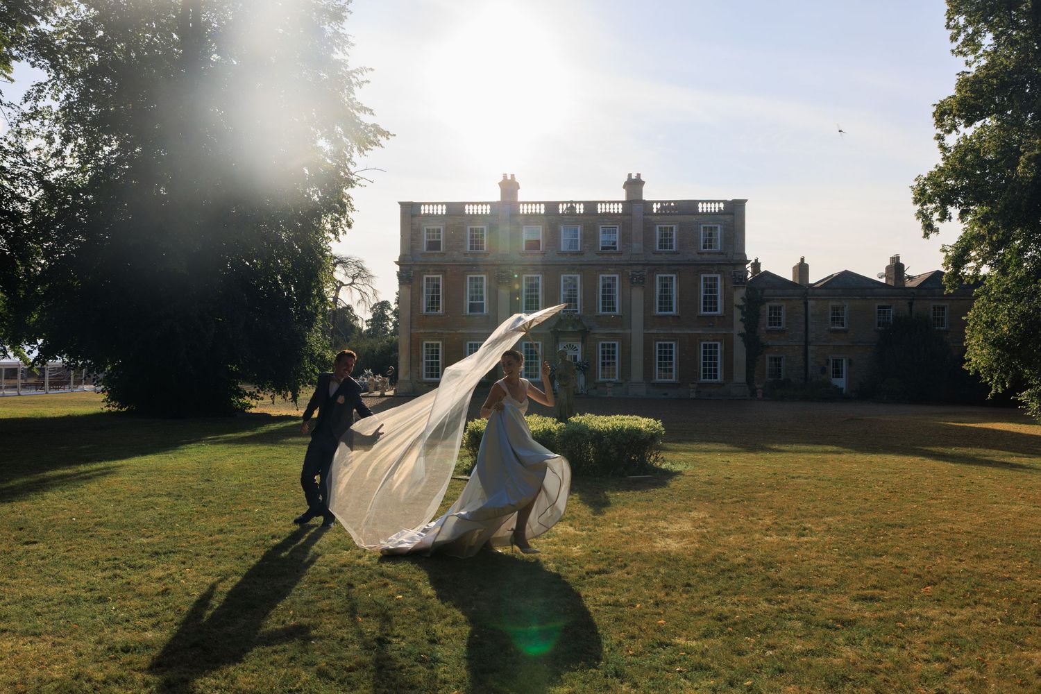 Newlyweds run joyfully across golden sunlit grounds of elegant red-brick Georgian manor, bride's veil billowing in wind.
