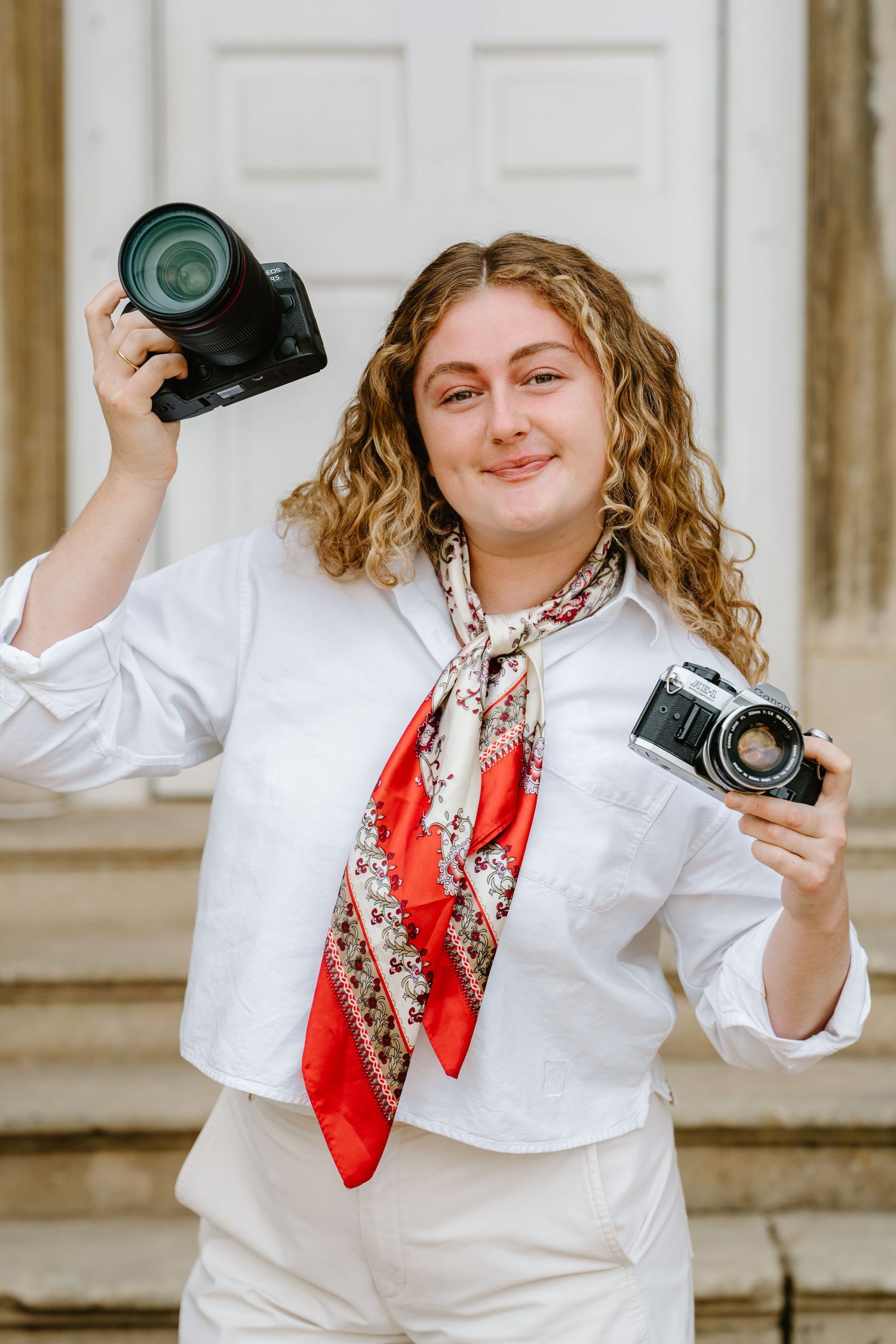 Smiling female photographer holds DSLR and vintage film camera, wearing white outfit with red floral scarf outdoors.