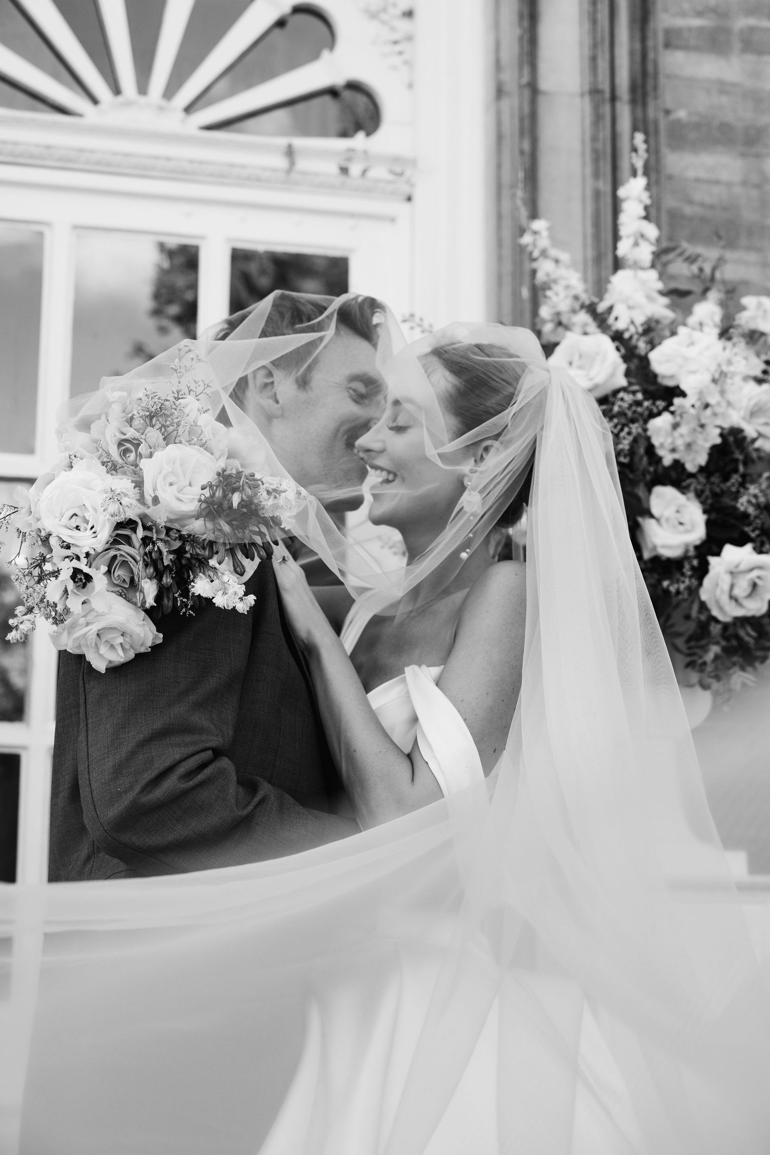 Black and white photo of bride and groom sharing romantic moment under flowing wedding veil.