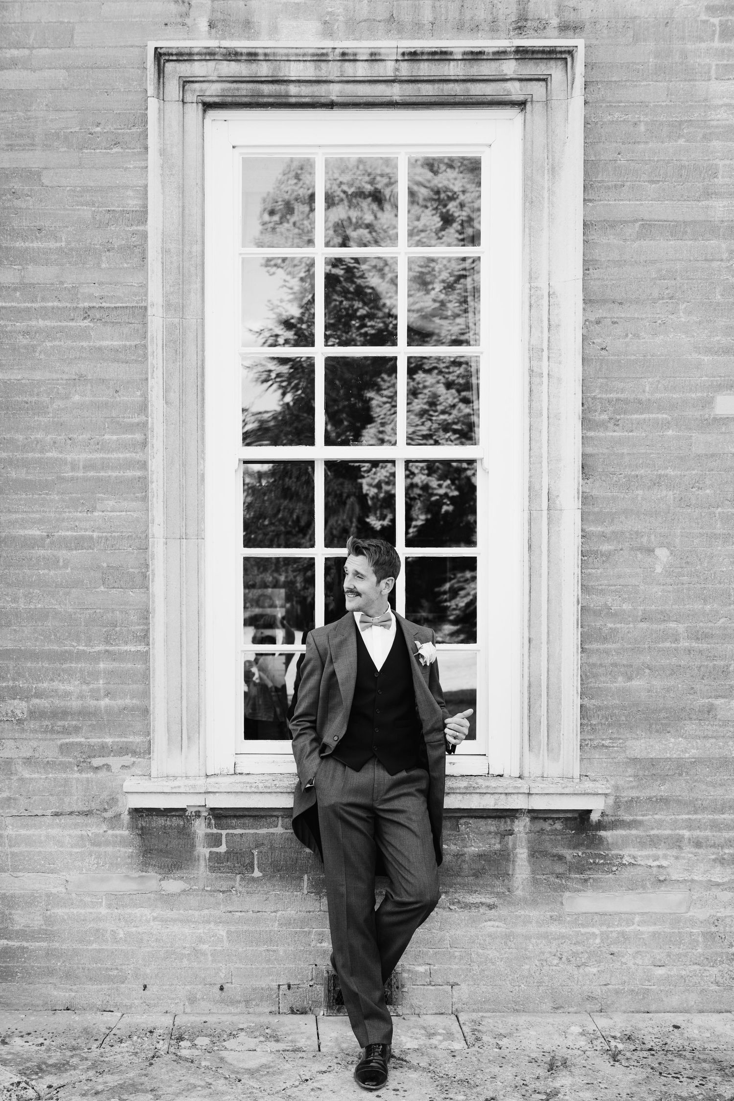 Dapper groom in suit leans against brick wall beside tall window in black and white portrait.