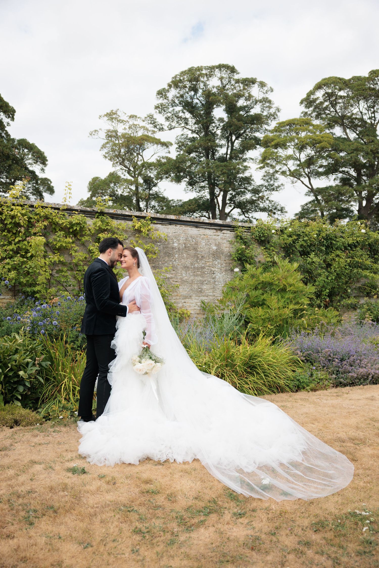 Bride in ruffled gown with cathedral veil and groom in black suit embrace in walled garden. Boughton House